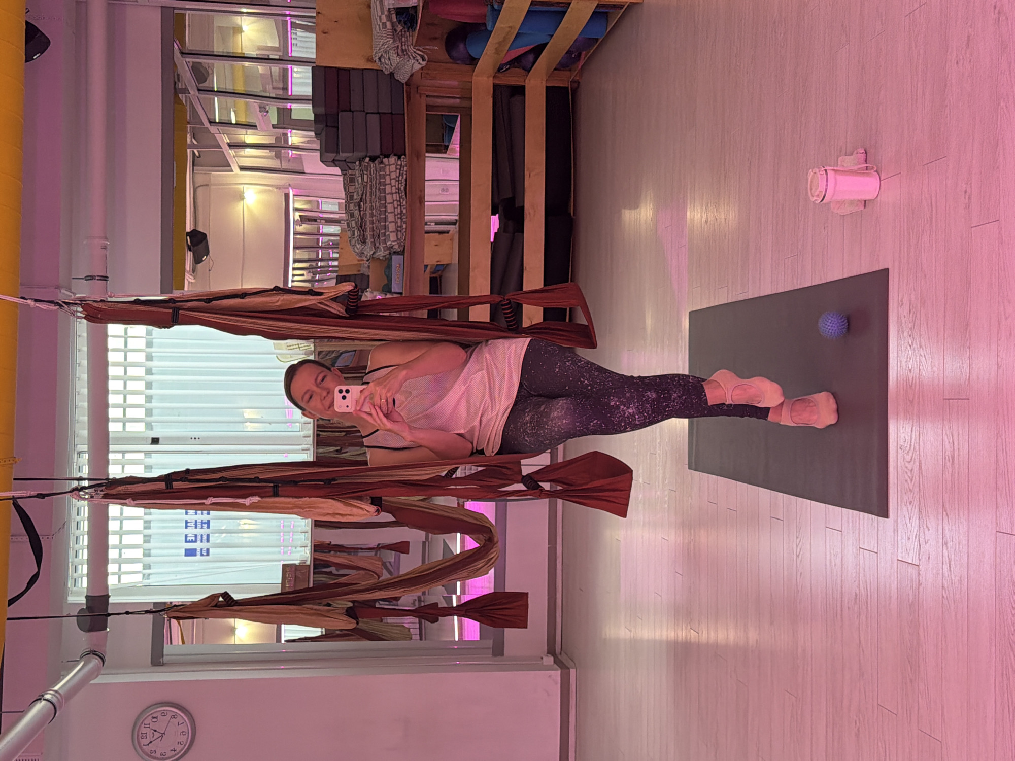 A woman is taking a selfie in an aerial yoga studio. She is holding a pink smartphone in her right hand and is positioned in the center of the frame, partially suspended in a yoga hammock. She is wearing a light-colored sleeveless top and patterned leggings, with pink ballet slippers on her feet. The studio has a light-colored wooden floor and is illuminated with pink lighting. In the background, there are yoga hammocks hanging from the ceiling, shelves stocked with yoga mats and blocks, and a clock on the wall. To the right of the woman, on the floor, are a blue massage ball and a pink water bottle with a towel. The setting appears to be a professional yoga or fitness studio.