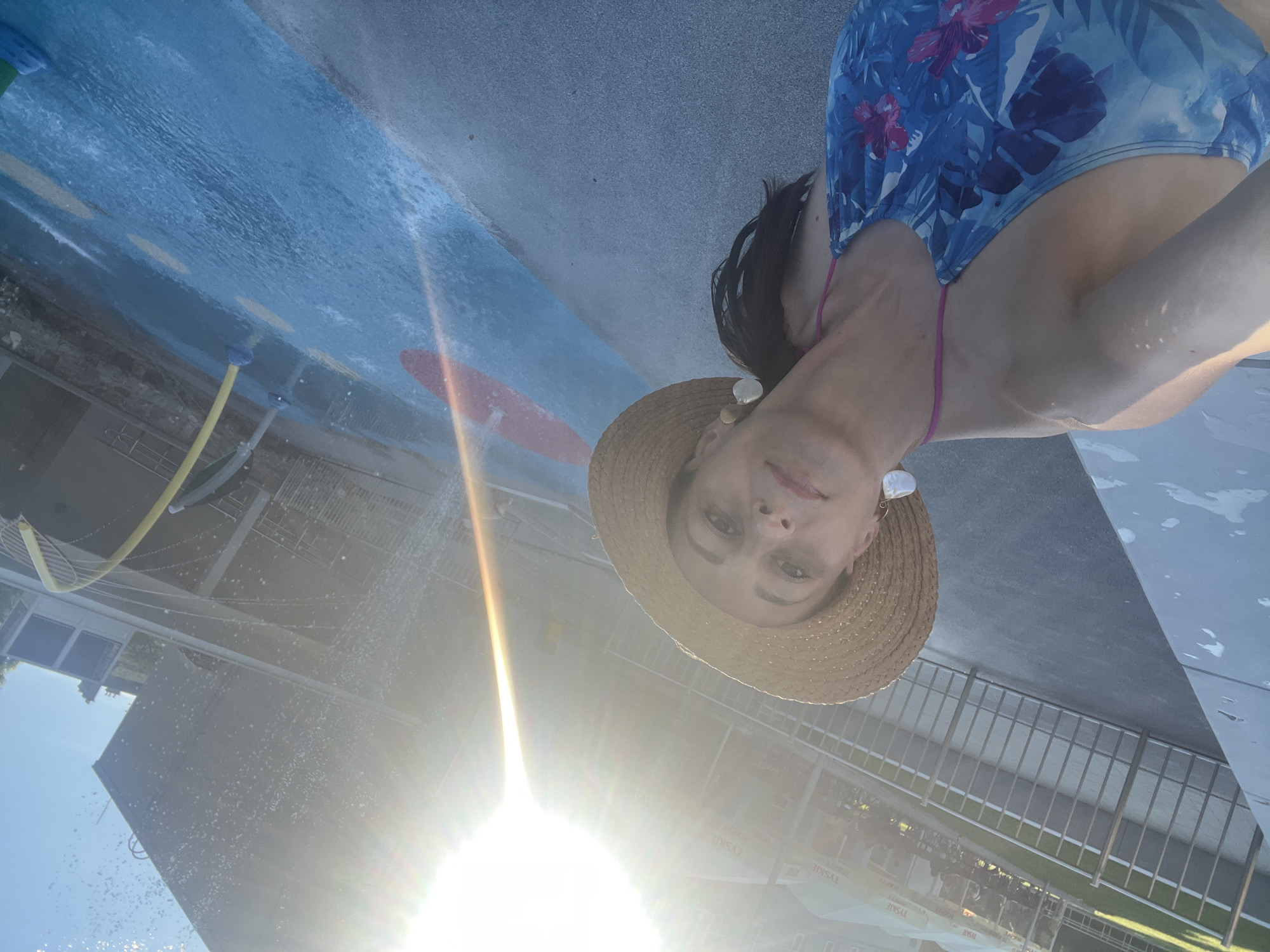 A woman wearing a blue floral swimsuit and a straw hat is taking a selfie outdoors. She is standing near a splash pad with water spraying in the background. The sun is shining brightly, creating lens flare. In the background, there are umbrellas and tables of an outdoor cafe. The overall environment appears to be a public outdoor recreational area in the city of Unknown, Unknown.