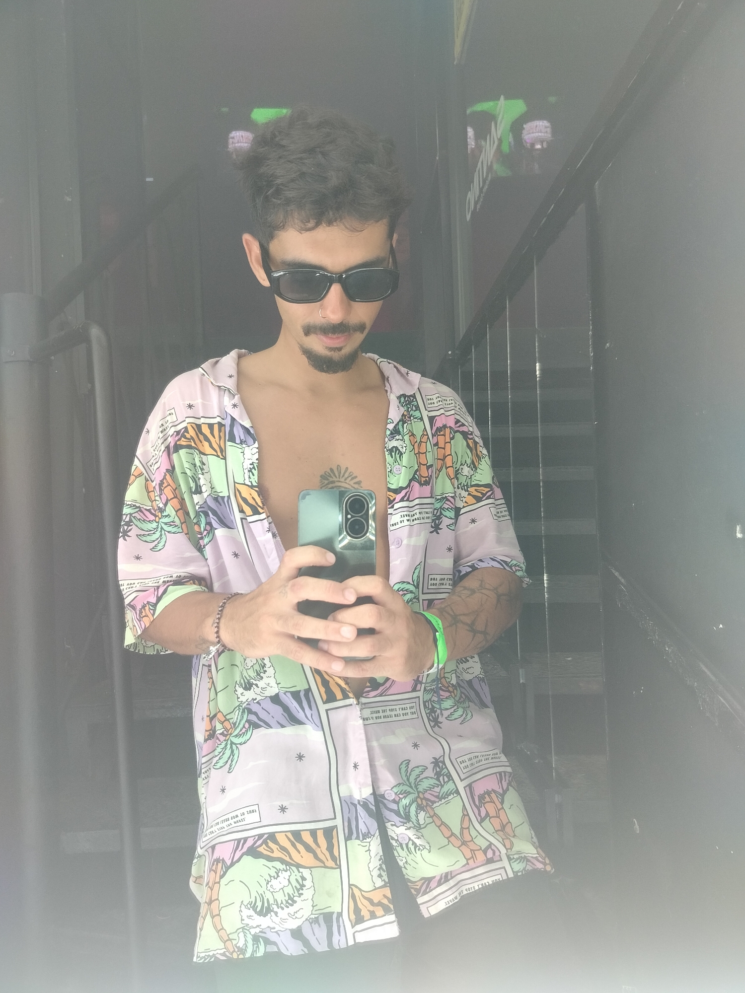 A man stands in front of a dark doorway and mirror with a staircase behind, taking a selfie. He is wearing sunglasses and a pink shirt with cartoons. The shirt is partially unbuttoned. He has a nose ring and a tattoo on his chest. He is holding a phone. The background is dark, but some signs with upside-down lettering are visible in the mirror. The location is likely Playa del Carmen, Mexico.