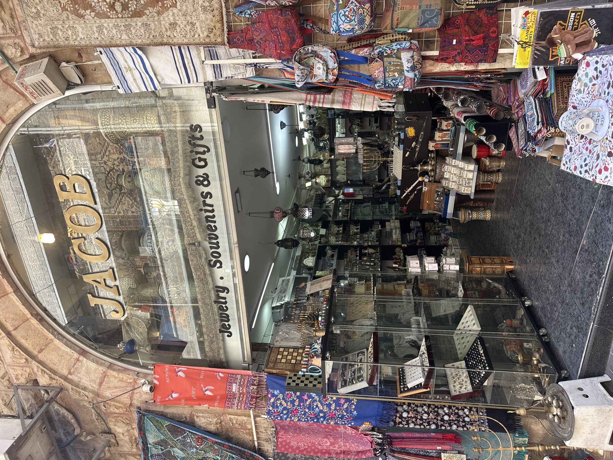 The image shows a storefront in Jerusalem, Israel, named "JACOB Jewelry, Souvenirs & Gifts." The shop is filled with various items, including jewelry displayed in glass cases, colorful scarves hanging outside, and several menorahs. Bags with intricate patterns are also visible. The shop's entrance is arched, and the storefront is decorated with a tapestry and a sign. The street outside is paved with dark tiles.