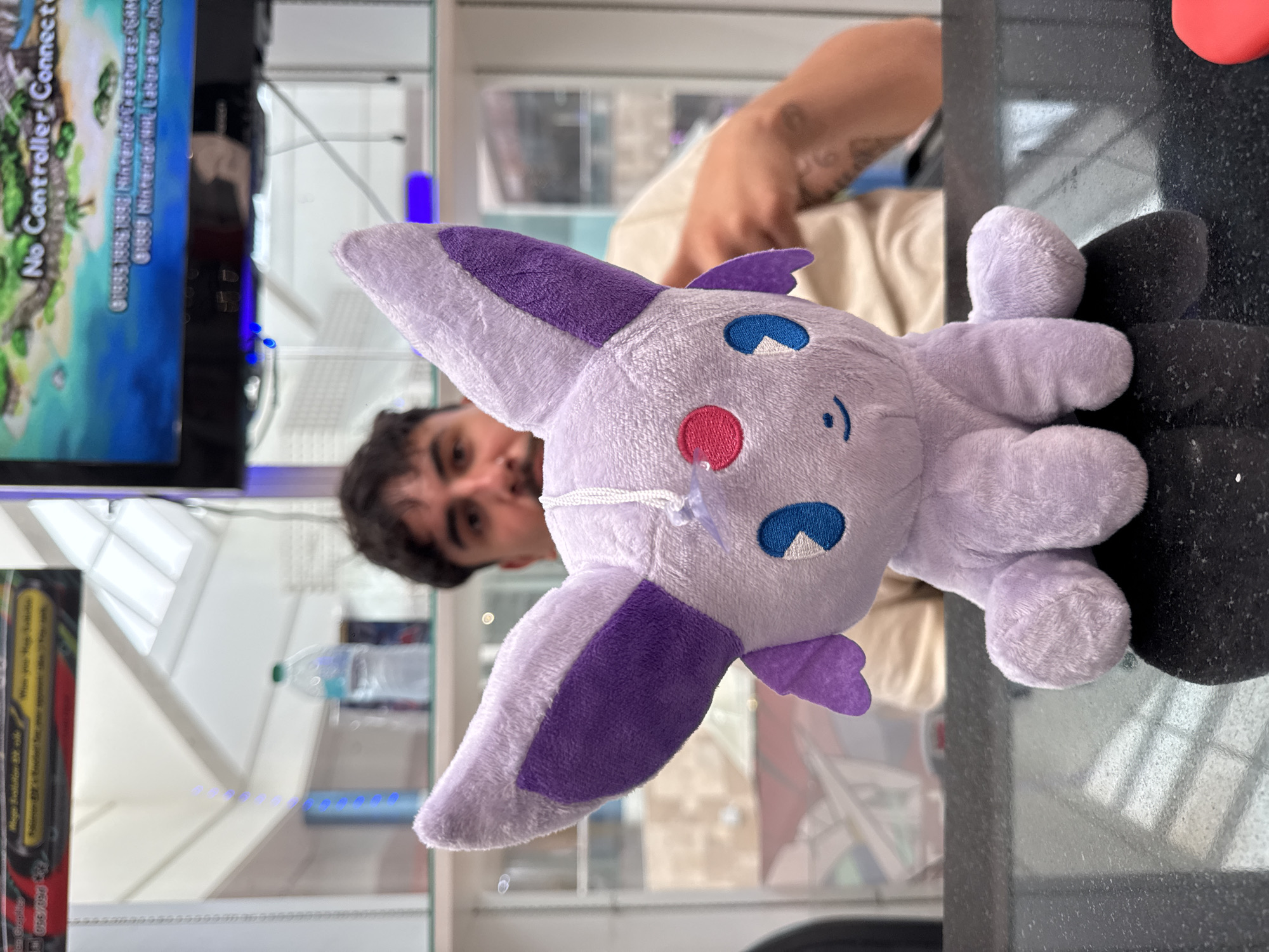 A close-up, eye-level, medium shot captures a light purple stuffed toy, identified as Espeon from Pokémon, positioned on a black reflective countertop. The plush toy faces the viewer, with its large ears, pink and blue eyes, and small smiling mouth clearly visible. A white string with a clear plastic tag is attached to the toy's head. In the background, a man with dark hair and a beard is partially visible, out of focus, wearing a beige t-shirt. He is seated behind the countertop. Further in the background, shelves are stocked with various items, including a clear plastic water bottle, and a television screen displaying a colorful image and text, possibly from a video game. The environment appears to be an indoor retail or entertainment establishment, with the presence of a television and shelves suggesting a store or arcade. The image was taken in El Paso, Texas, United States of America.