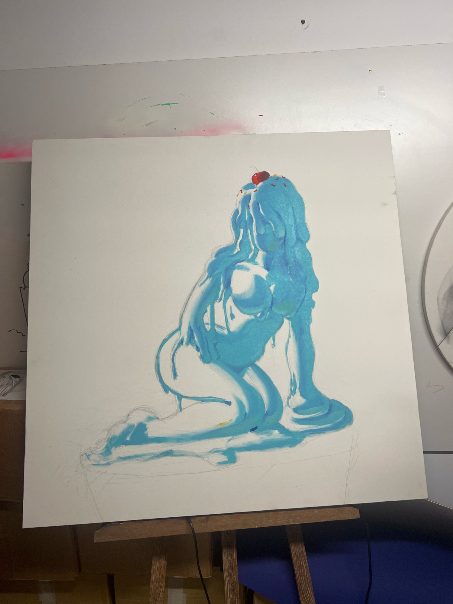 The image shows an artist's studio with a large canvas on an easel. The canvas features a painting of a stylized, dripping blue figure that resembles melting ice cream or slime, topped with a cherry and sprinkles. The figure is in a kneeling or crouching pose. Faint pencil sketches are visible beneath the painted areas, suggesting the painting is in progress. The studio environment appears to be a well-lit room with white walls. Some faint marks of pink and green spray paint are visible on the wall in the upper left quadrant of the image. There are also some tools and other artistic materials visible in the background, including what appears to be another drawing on the left and a circular sketch on the right. The scene is domestic and creative. There is no indication of the location being El Jadida, Morocco.