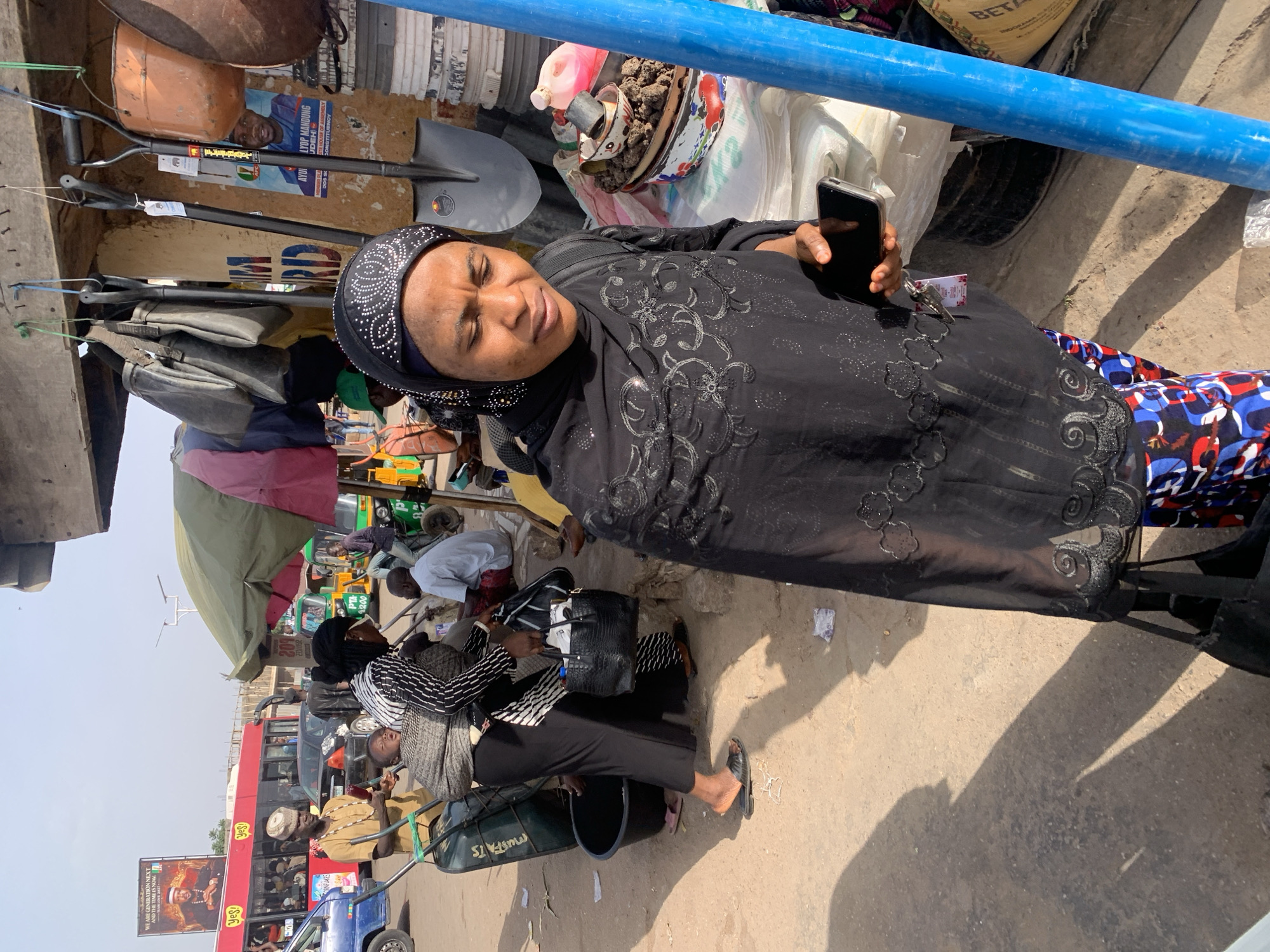 This image captures a lively outdoor market or street scene in Jos, Nigeria, under bright, clear daylight, indicated by strong shadows. In the foreground, a woman wearing a black headscarf with white patterned embellishments and a matching long-sleeved tunic with intricate grey embroidery holds a black smartphone. She looks towards the viewer with a neutral expression.
Around her, the environment is bustling with various objects and activities. A prominent blue pipe runs horizontally across the upper mid-ground. Nearby, several shovels hang, an orange bucket is visible, and another container holds soil or a similar substance. A black handbag sits beside the woman, and a green wheelbarrow with "MUSFATS" printed on it is partially visible.
In the background, numerous individuals are engaged in daily market activities; some sit, others walk, and vendors attend to their stalls. Makeshift market structures with pink and olive-green awnings line the street. Traffic includes a large red double-decker bus, smaller vehicles, and green auto-rickshaws (Keke Napep), reinforcing the busy urban atmosphere.
Visible text includes political campaign messages such as "AYUI LYOP MANDUNG," "UDEH," "TE HOUSE OF ASSEMBLY," and "CONSTITUENCY." Other signs read "WE ARE GENERATION NEXT AND THE TIME IS NOW.," "Yes!" (repeated), "CORNFLAKES," "PTA 2200," "BETA" (on a sack), "SRD," and "SOC." The scene portrays a vibrant and typical day in a Nigerian marketplace.