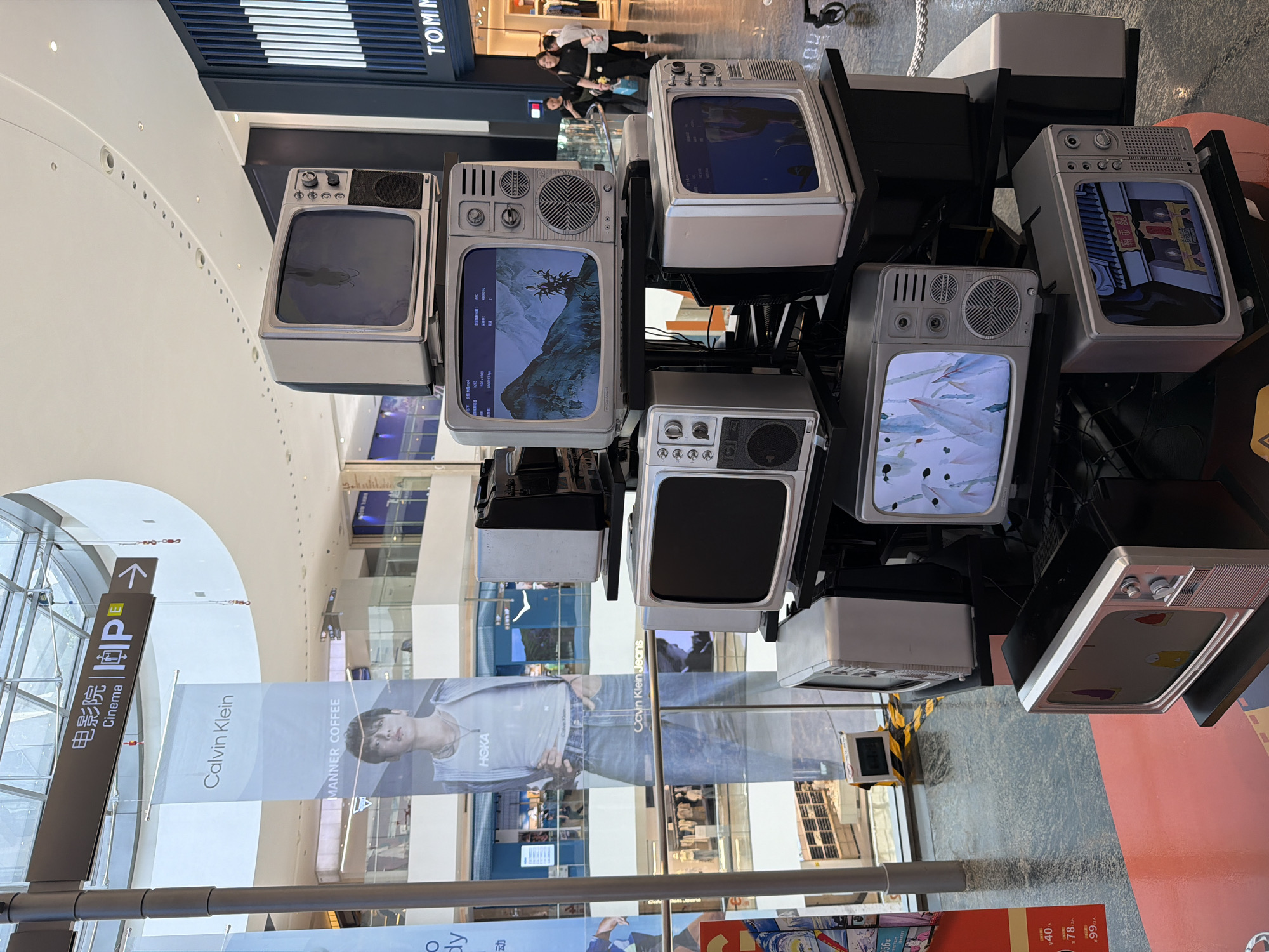 In the Yuzhong District of China, within what appears to be a multi-level shopping mall, a tower of vintage-style televisions is displayed as an art installation or promotional display. The televisions, varying in size and model, are stacked precariously on black metal shelves. Some screens display digital content, including abstract art and what seem to be video game graphics. The background reveals the mall's interior, with signage for "Cinema" and various stores, including a "Calvin Klein" banner and the facade of a "TOMMY" store. Several individuals are visible in the distance, walking through the mall. The flooring is a polished, reflective surface, and overhead lighting illuminates the scene.