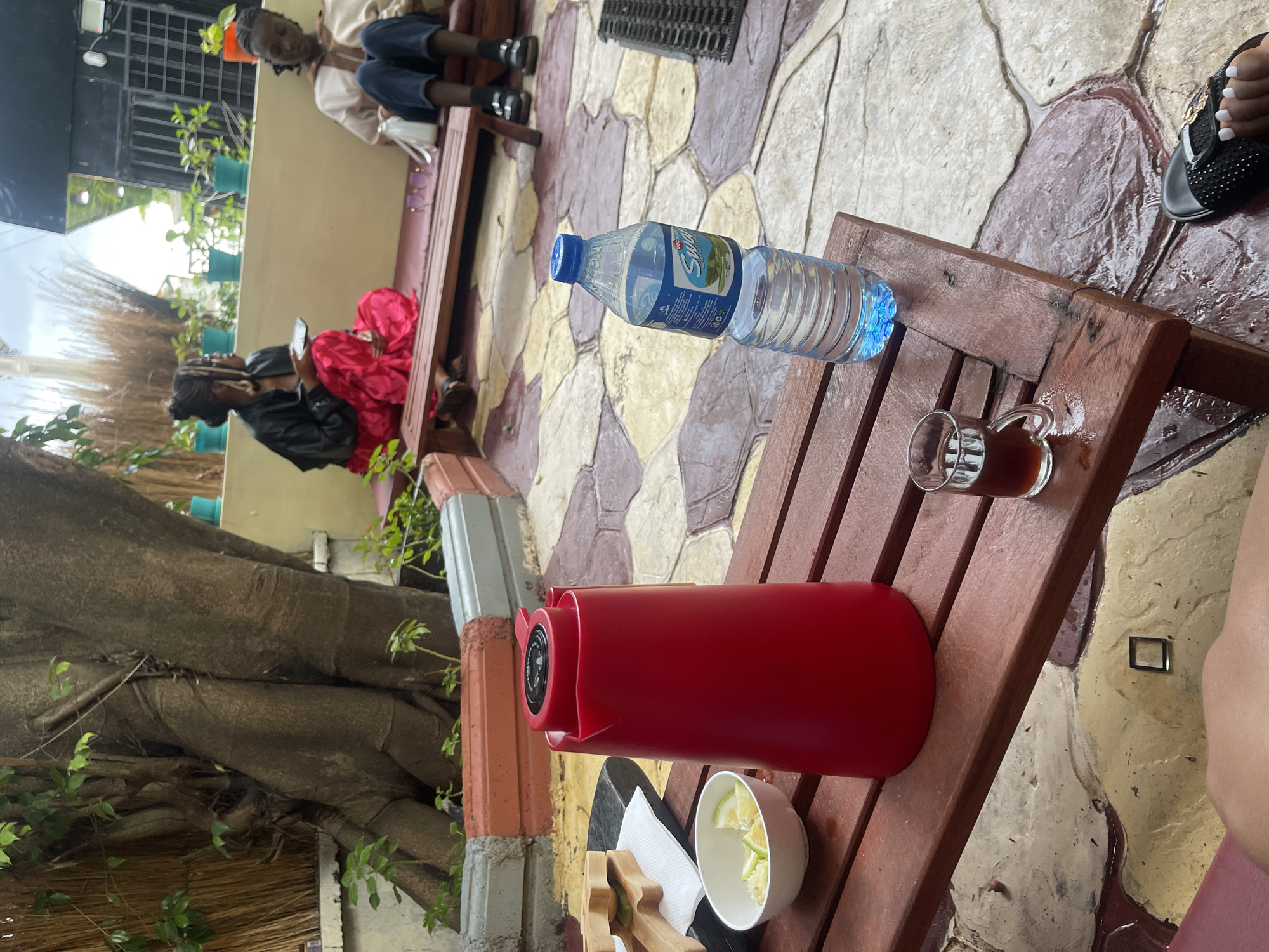 The image depicts an outdoor seating area in Gura Top, Nigeria. In the foreground, a red thermos sits on a wooden table, next to a bowl of sliced lemons and a small glass of dark liquid. A partially visible water bottle is also on the table. Two women are seated on a bench in the background. One, wearing a black jacket and a bright pink skirt, holds a phone. The other, in a light-colored shirt and dark trousers, appears to be looking down. A large tree trunk is visible on the left side of the frame, and potted plants line a wall behind the bench. The ground is paved with irregularly shaped stones.