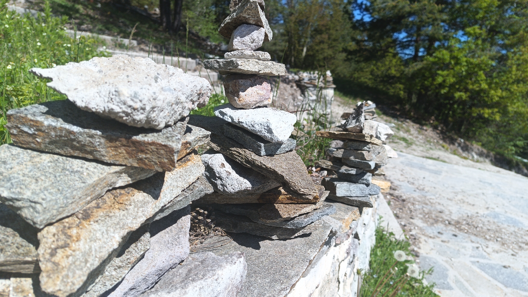 This image depicts a stone wall in Sini Vrah, Bulgaria. There are three stone cairns built on top of the wall. The cairn closest to the viewer is made of rough-cut rectangular stones. The middle cairn is composed of flat stones and round rocks stacked atop each other. The third cairn in the background is built of smaller flat stones. The ground behind the stone wall is a paved road leading uphill into a wooded area with trees visible. To the left of the wall, there is a patch of green vegetation. The sky is blue. The image is taken during daylight.