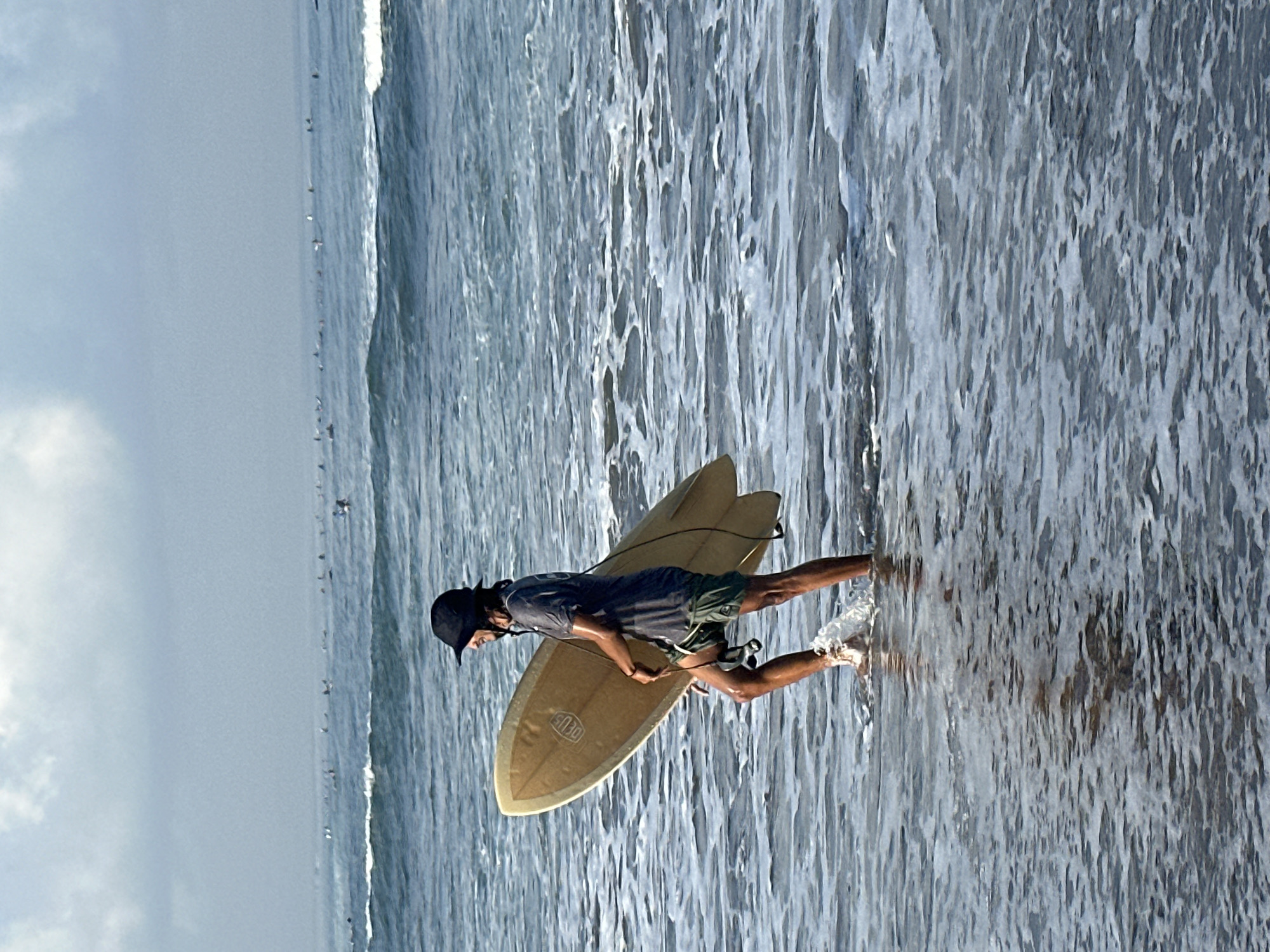 A lone surfer wearing a dark bucket hat, a grey t-shirt, and green shorts walks into the ocean carrying a tan surfboard on their right side. The surfer is in profile, facing left, with their left leg submerged up to the knee in the water, kicking up a splash. The water is calm and shallow around the surfer, with gentle waves breaking in the background. The ocean stretches out to a hazy horizon where numerous other surfers are visible. The sky is overcast, with soft clouds. The scene is set in the waters off the coast of North Kuta, Indonesia, known for its surf breaks.
