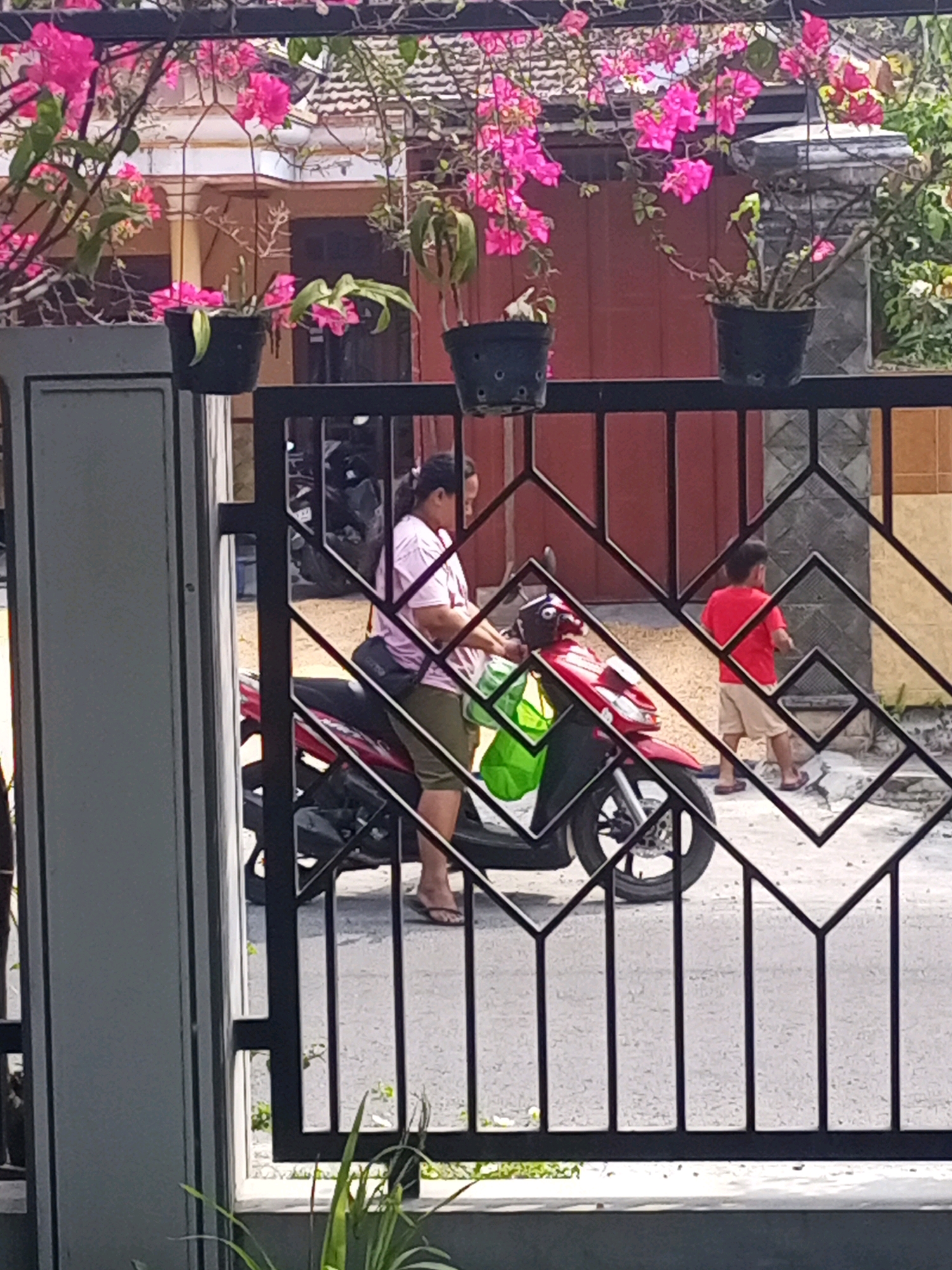 The image shows a street scene in Ngasem, Indonesia. In the foreground, a black metal fence with a geometric pattern obscures part of the view. Beyond the fence, a woman wearing a pink shirt and olive green shorts is standing next to a red and black motorcycle. She has a dark-colored bag slung over her shoulder and is holding a green bag in her left hand. To the right of the motorcycle, a young boy in a red t-shirt and light brown shorts walks away from the camera, wearing sandals. The background features a building with a red tiled roof and a tan-colored wall, partially covered by pink bougainvillea flowers cascading from hanging planters. The ground appears to be paved with asphalt.