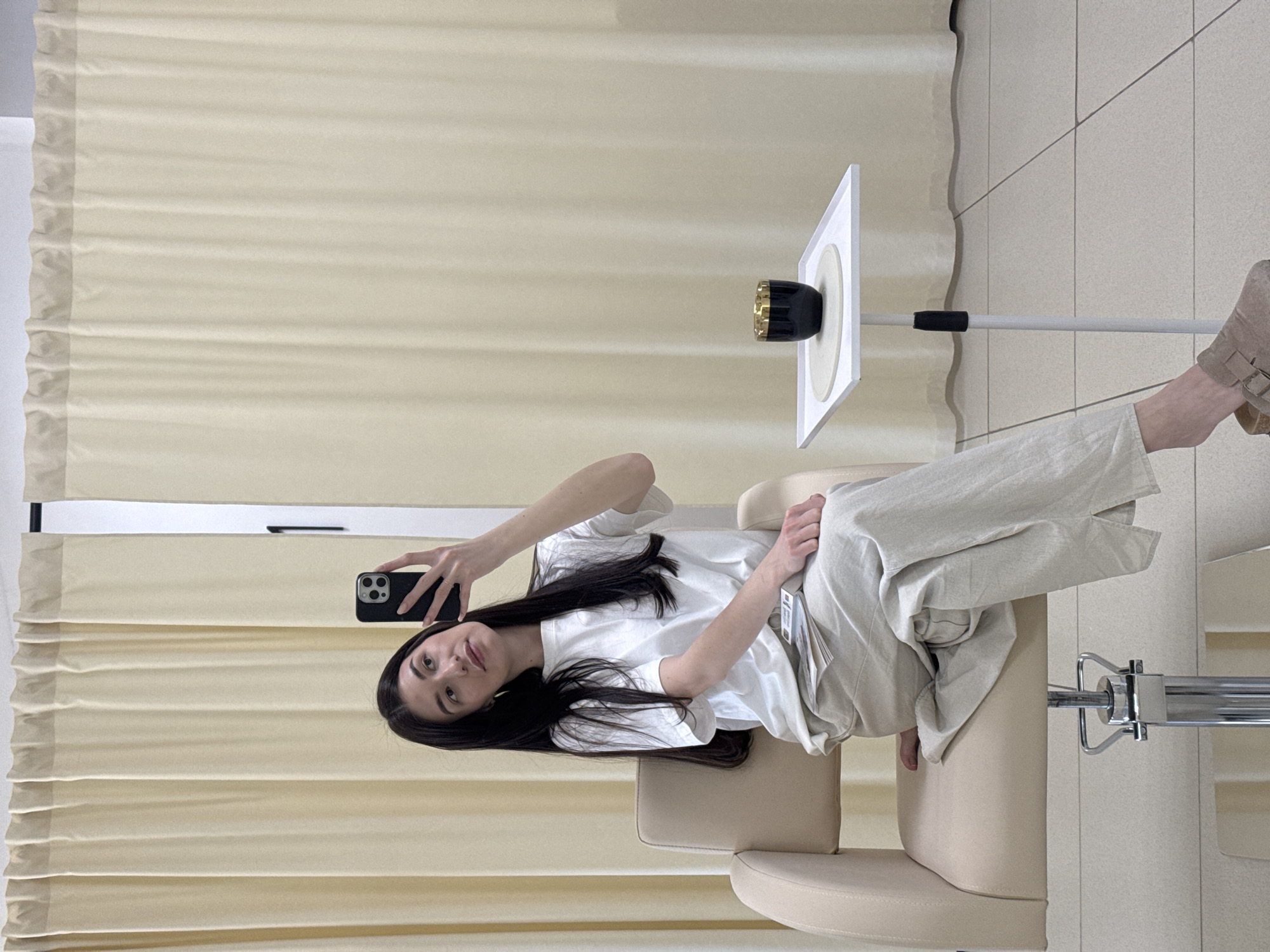 A single female subject is seated in a beige salon-style chair within an indoor setting. She has long, dark hair and is wearing a white t-shirt, light beige wide-leg pants, and a light beige buckled slip-on on her left foot. Her right hand holds a black smartphone, positioned upwards and pointed towards herself, indicating a self-portrait or mirror capture. A stack of small, white rectangular papers or cards rests on her left thigh. To her right, a white, slender side table supports a black cup with a gold rim, resting on a white circular coaster. The background features light cream-colored, pleated curtains extending from ceiling to floor. The floor is covered in light-colored square tiles. The environment suggests a beauty salon or studio. No specific visual cues within the image identify the location as Chaikovsky, Russia.