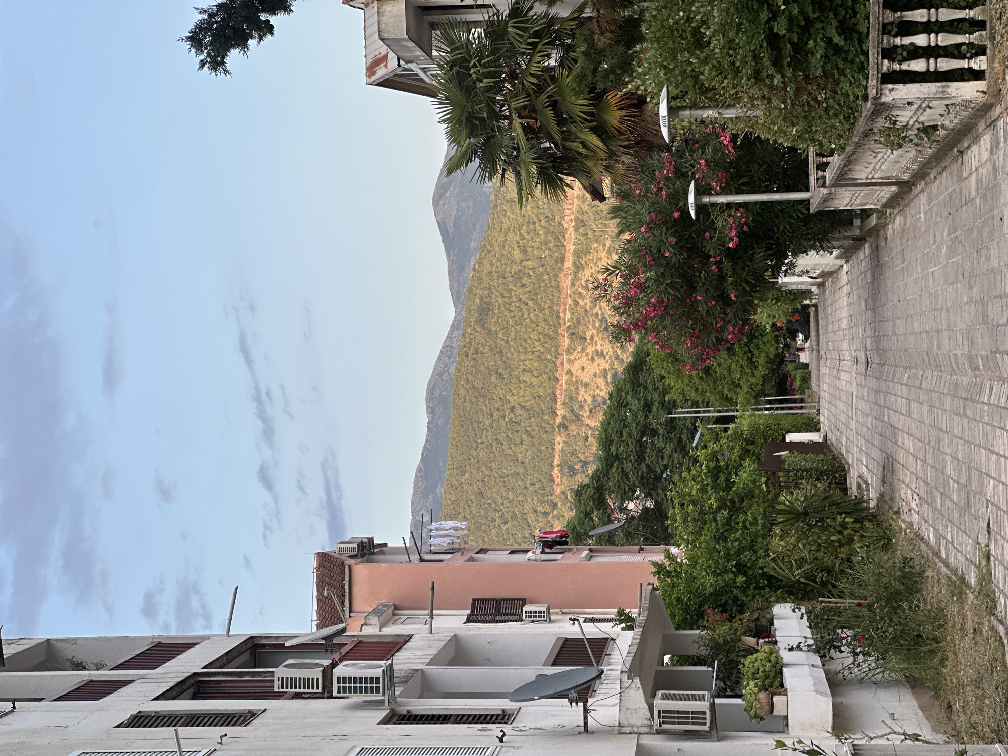 The image depicts a street scene in Budva, Montenegro. On the left, a white apartment building features balconies with red railings and visible air conditioning units and satellite dishes. Adjacent to it is a pink building with laundry hanging on a clothesline. The street itself is paved with cobblestones and lined with greenery, including flowering bushes and trees. In the background, rolling hills covered in vegetation rise towards a clear sky with scattered clouds. Two modern streetlights are visible along the right side of the path. The overall impression is a residential area with a view of natural landscape.