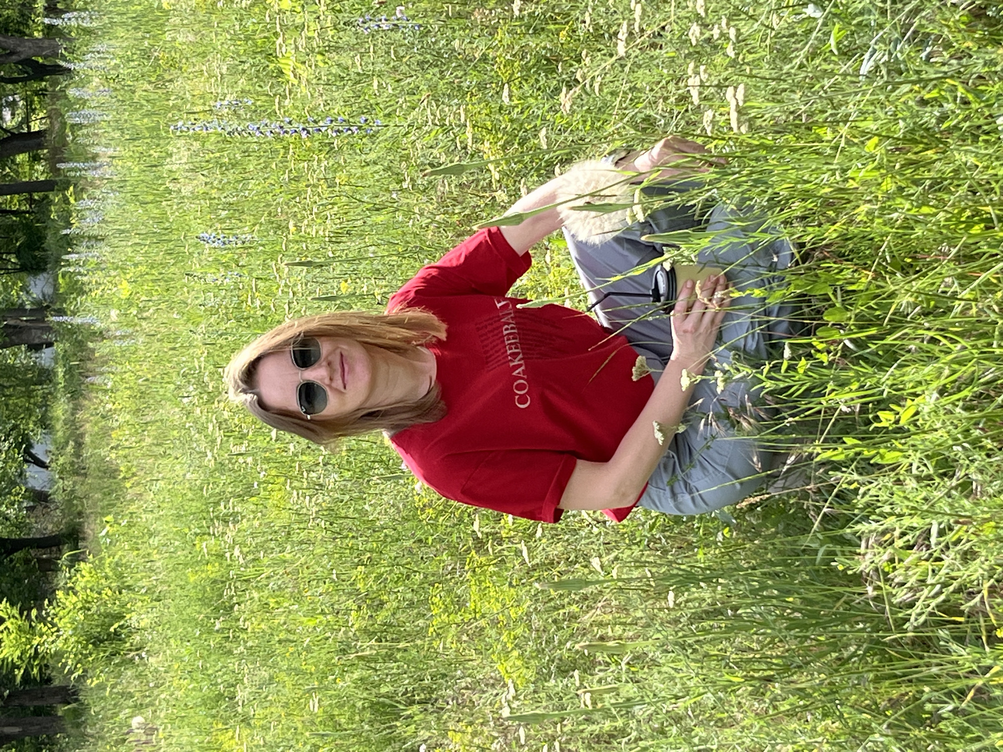 A woman is crouching in a field of tall green grass and wildflowers, with trees in the background. She is wearing sunglasses, a red t-shirt with white text that reads "COAKEEBALL", and grey pants. She is holding a disposable cup in her right hand. The image is taken outdoors on a sunny day. The specific location within the city of Kryvyi Rih, Ukraine cannot be determined from the image alone.