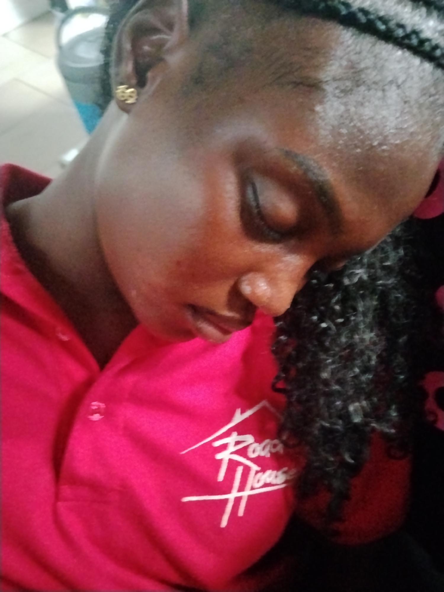 The image is a close-up, high-angle shot of a young Black woman. She is wearing a bright pink polo shirt with a white logo on the left side of the chest. The logo appears to be text that reads "Raga House." Her eyes are closed, and she is looking downwards. Her hair is styled in braids and falls over her right shoulder. She is wearing a gold earring in her left ear, which appears to be shaped like the number 69. The background is blurred and out of focus, but it appears to be an indoor setting with some light-colored elements and possibly a blue container in the upper left quadrant. The image is lit from above, creating highlights on her forehead and cheek. The woman is positioned slightly to the left of the center of the frame. The image does not provide any information about the city of Howayi, Nigeria.
