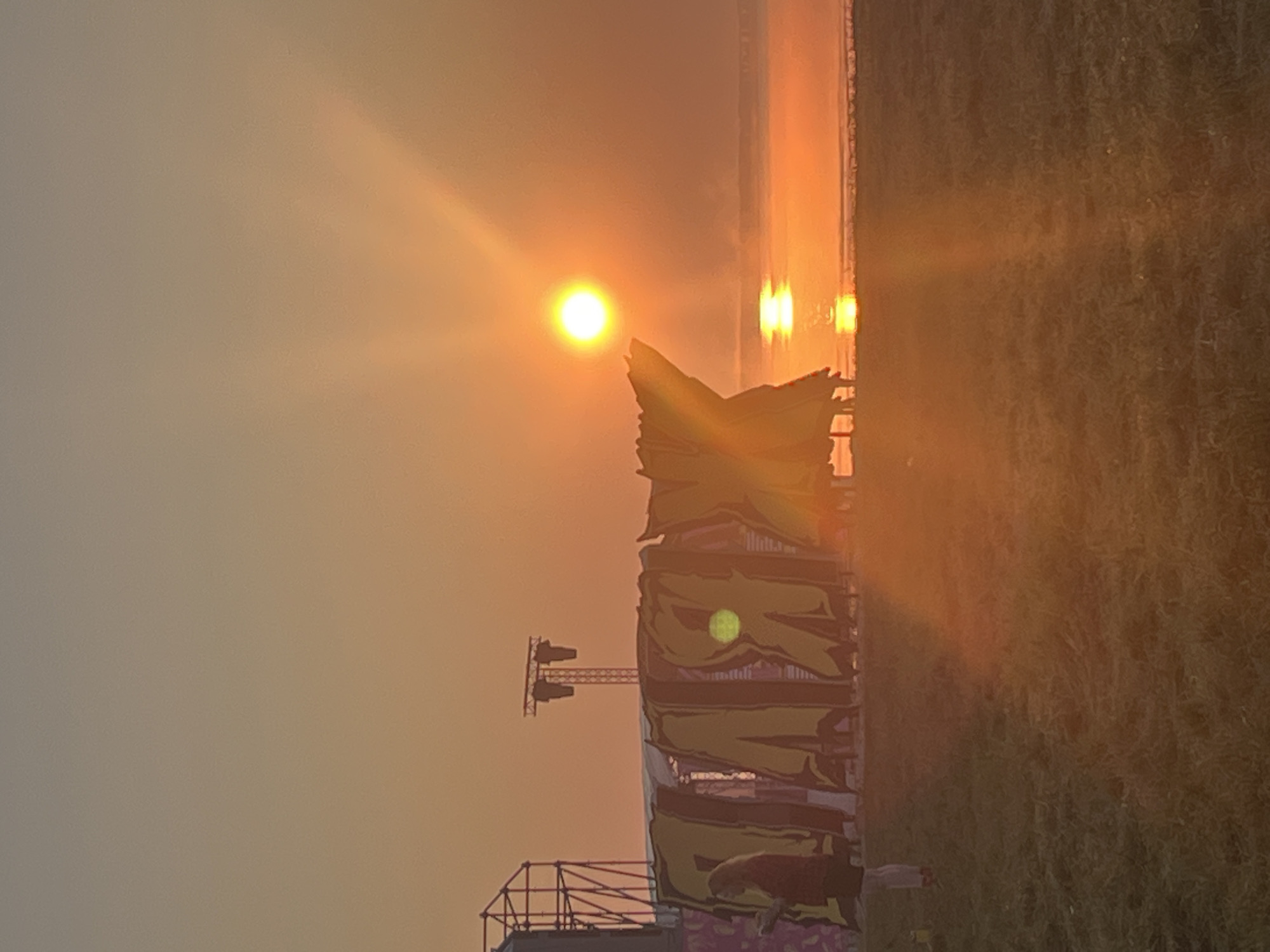 An outdoor festival stage is visible in the foreground at sunrise, with a hazy sun low on the horizon reflecting on the water. A person stands to the left, facing the stage. The stage is decorated with large, colorful banners that appear to be made of fabric. In the background, a large metal structure, possibly scaffolding or part of a larger stage setup, is visible. The sky is a gradient of orange and yellow hues, typical of a sunrise. The ground in the foreground is covered in green grass. The location is identified as Surakovo, Russia.