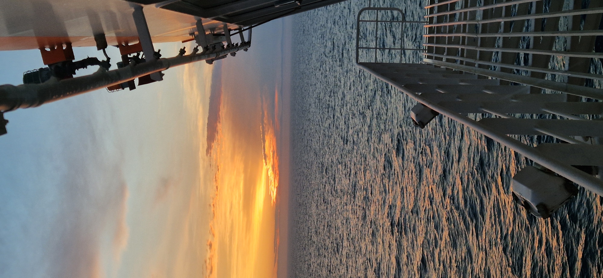 A vertical, low-angle shot captures a sunset over the ocean from the deck of a vessel. The sky transitions from a pale blue at the top to a vibrant orange and yellow as it nears the horizon, with wispy clouds scattered throughout. The sun is a bright, diffused glow peeking through the clouds just above the horizon. The ocean below is a deep blue, with small waves rippling across its surface, reflecting the warm hues of the sunset. In the foreground, the side of a ship is visible, with a white metal railing and staircase leading upwards. Several industrial-looking fixtures, possibly lights or sensors, are mounted on the ship's exterior. The scene conveys a sense of peaceful travel or observation at sea during twilight.