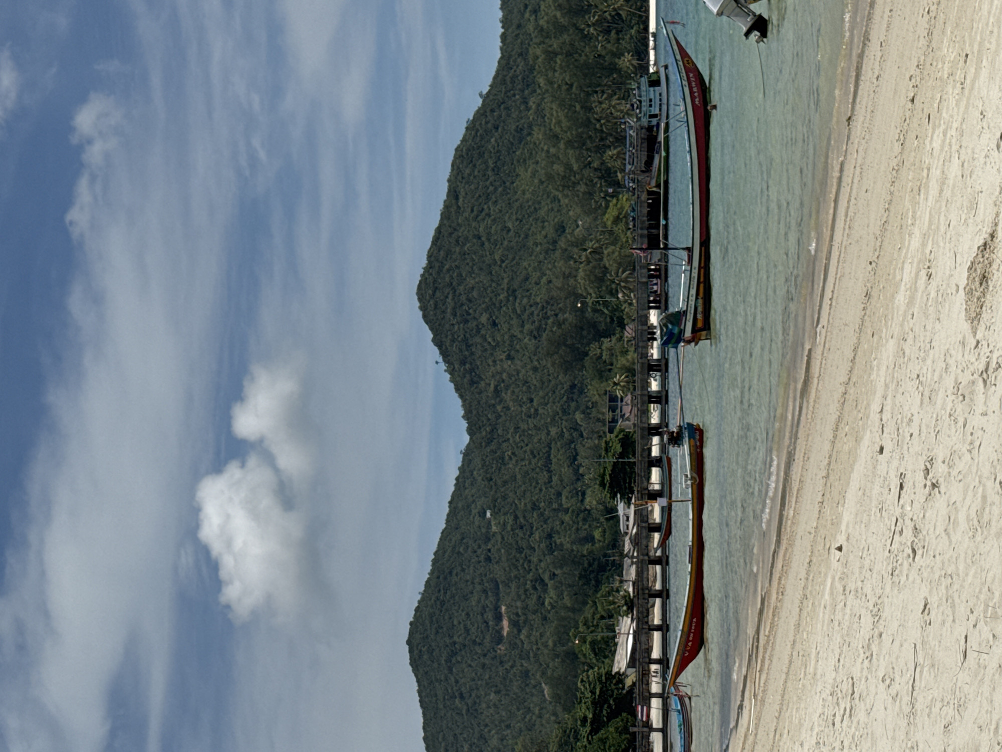 The image depicts a beach scene in Baan Chaloklum, Thailand, featuring a white sandy beach, clear turquoise water, and a lush green, mountainous backdrop. Several longtail boats are moored along a wooden pier that extends into the water. The boats are colorful, with red, blue, and yellow accents. The sky is blue with scattered white clouds. The scene appears to be taken during the day, under sunny conditions.