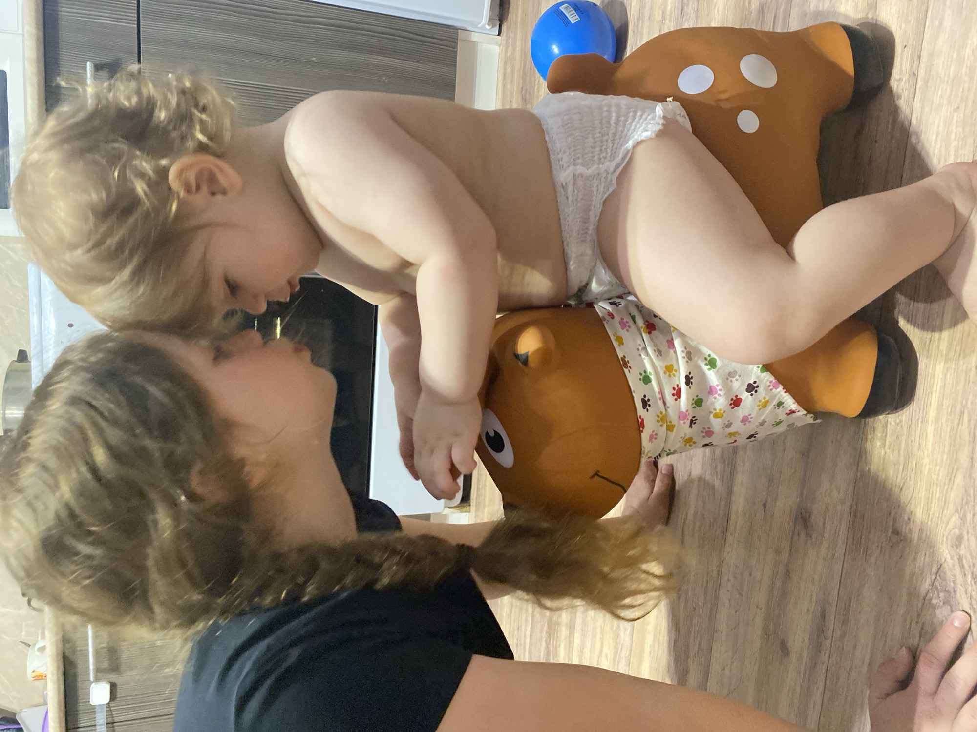 A young child in a diaper is on top of a brown inflatable toy shaped like an animal, possibly a deer. The child faces left and appears to be interacting with an adult woman who is kneeling or sitting. The woman is in a black shirt. The environment is indoors on a wood floor.