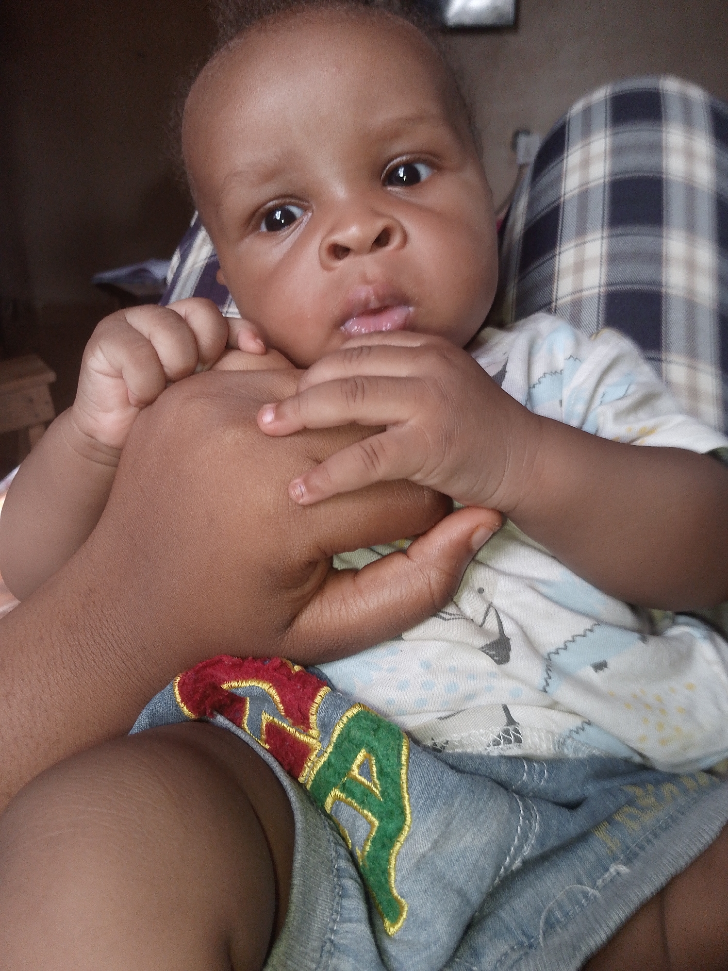 A baby with dark skin is being held by an adult in an indoor environment. The baby is wearing a light-colored shirt with a printed design and denim shorts with colorful embroidered patches. The adult's hand is visible, with the baby holding onto it. The background includes a plaid fabric surface and a glimpse of a wooden object. The location is in an unknown city.