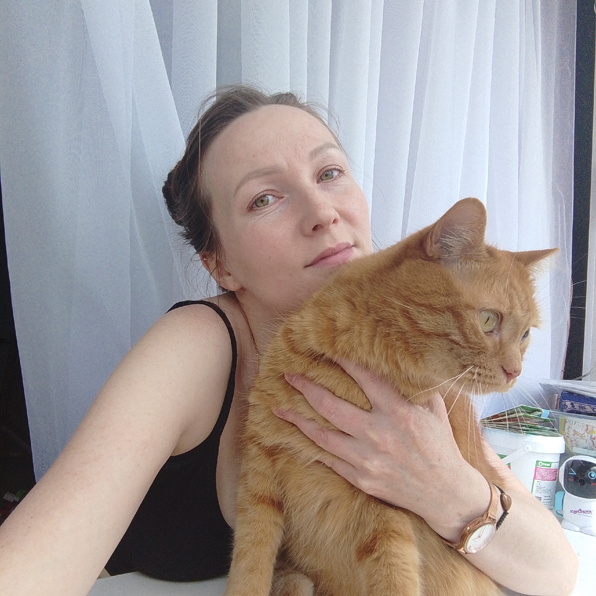 A casual portrait features a woman holding an orange tabby cat indoors. The woman, with light brown hair pulled back, looks directly towards the camera with a calm expression, her green eyes visible. She wears a black tank top and a rose gold watch on her left wrist. Her right arm is wrapped around the cat.
The ginger cat is positioned in front of her, facing right and looking slightly downward, its ears perked. Its fur appears thick and well-groomed.
The background consists primarily of sheer white curtains or blinds, allowing diffuse light into the scene, suggesting daytime. On a surface to the right of the cat, several objects are visible, including a white bucket with text "E-Prese" and "13." printed on it, and a small white robot-like figure with blue accents. Cyrillic text "Юный Я" (Yunyy Ya) is partially visible on the robot figure. The overall setting appears to be a bright, domestic space.