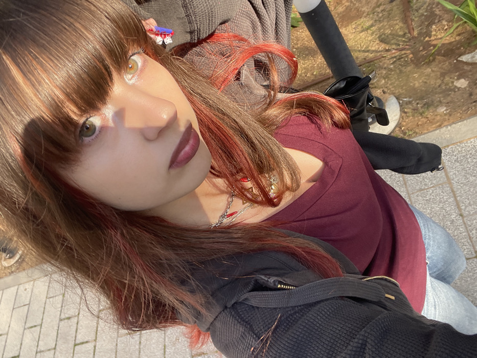 An outdoor, high-angle portrait captures a female individual from the chest up, positioned on a paved walkway in Raanana, Israel. Her long, medium brown hair features red highlights, with bangs framing her face. She wears dark lipstick. Her right eye is green/hazel, and her left eye is lighter, golden/hazel. Sunlight illuminates the left side of her face while casting a shadow across her nose. She is dressed in a maroon V-neck shirt, an open dark gray textured hoodie, and light blue denim jeans. Accessories include a silver chain necklace with multiple small red chili pepper charms, an obscured gold-toned circular pendant, and a multi-component earring on her left ear featuring white dice-like elements and a blue rectangular piece. The ground comprises light-colored pavers on the left and a strip of dirt and sparse grass on the right. A dark, cylindrical object is visible on the far right edge of the frame.