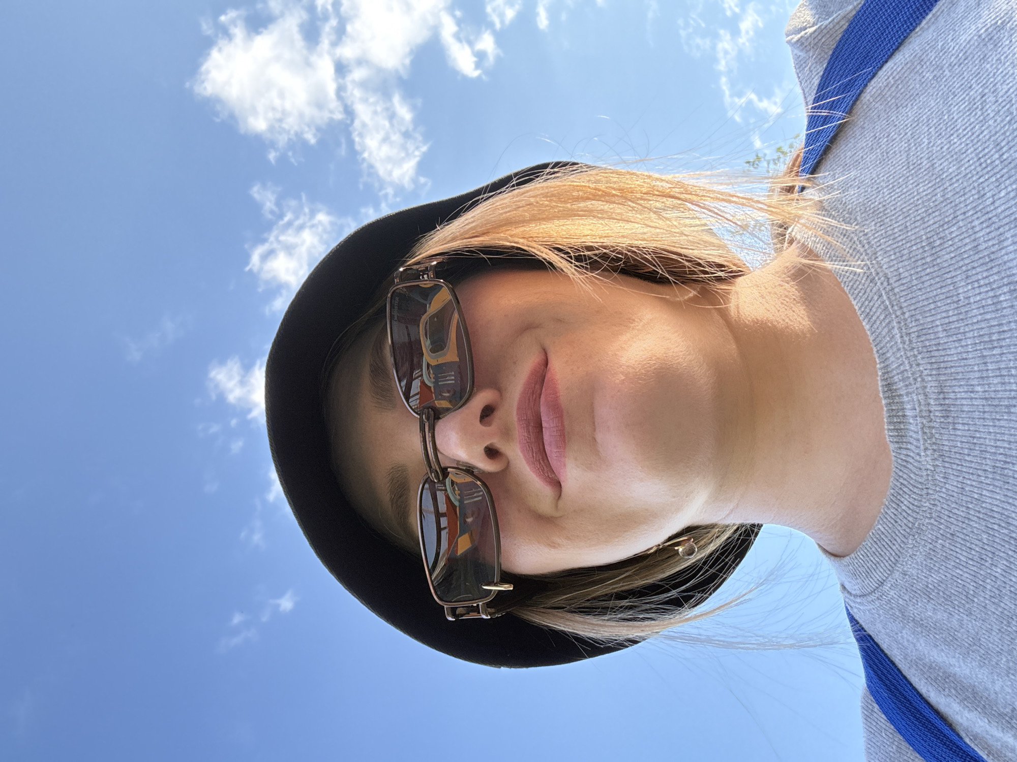 A low-angle, close-up, outdoor, waist-up selfie shows a young woman wearing a black bucket hat and sunglasses. The woman has blonde hair and is wearing a gray sweatshirt with a blue strap over her shoulder. She is smiling and looking up at the sky, which is blue with scattered clouds. The image appears to be taken in a park or other outdoor setting, possibly in Kazan, Russia. The reflection in her sunglasses shows what appears to be a street scene.