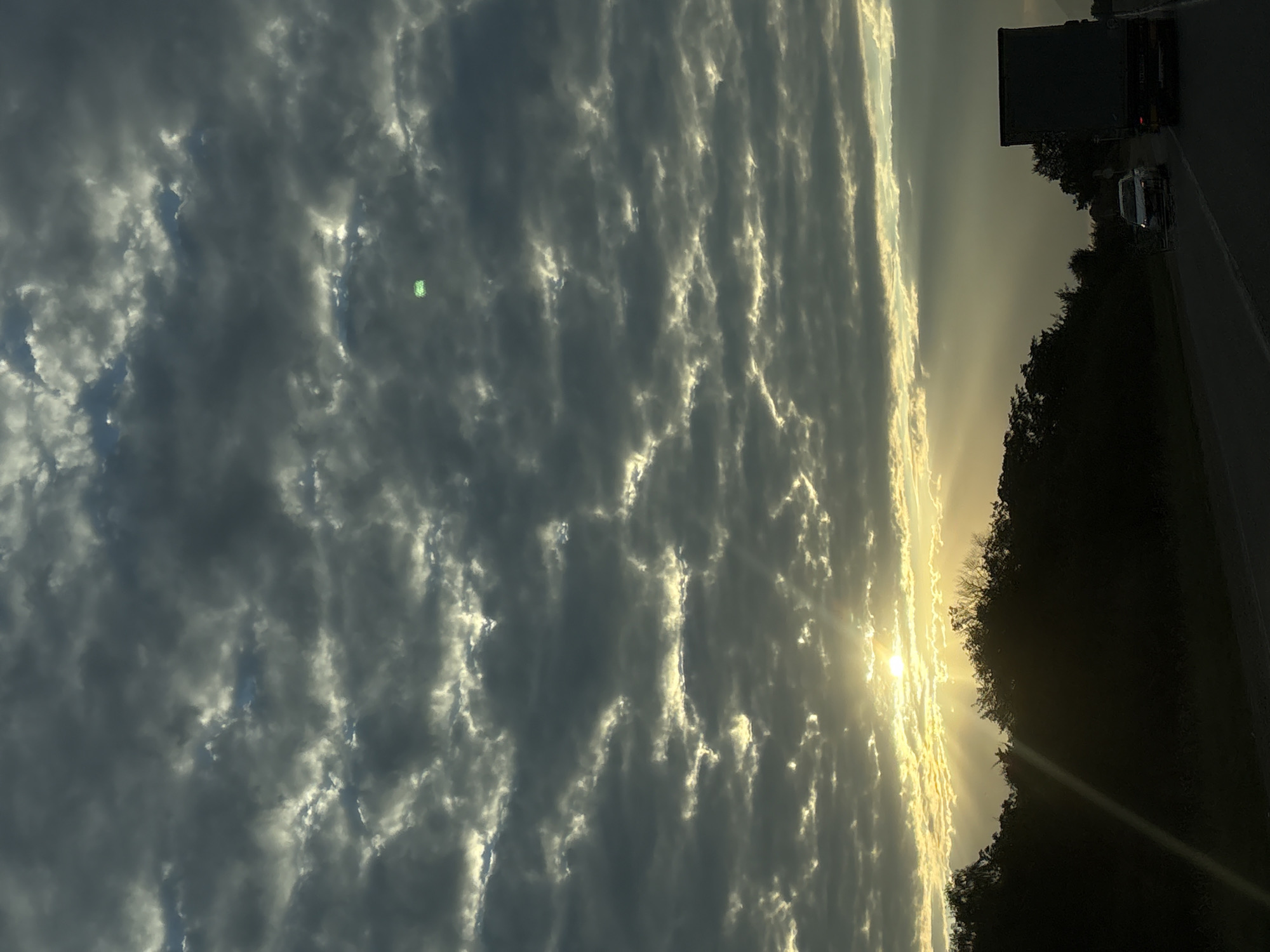 The image depicts a rural highway scene in Lysogorskaya, Russia, under a dramatic sky. The sun is low on the horizon, casting long shadows and creating visible sunbeams that pierce through textured, dark clouds. The light illuminates the edges of the clouds, giving them a luminous quality. A dark line of trees runs along the side of the road, silhouetted against the brighter sky. Two vehicles are visible on the asphalt road: a white passenger car in the left lane and a dark-colored truck in the right lane, traveling in the same direction. The road appears to be a two-lane highway. The overall atmosphere is one of dusk or dawn, with the natural landscape dominating the scene. A bright green speck is visible in the upper portion of the sky, likely an artifact.
