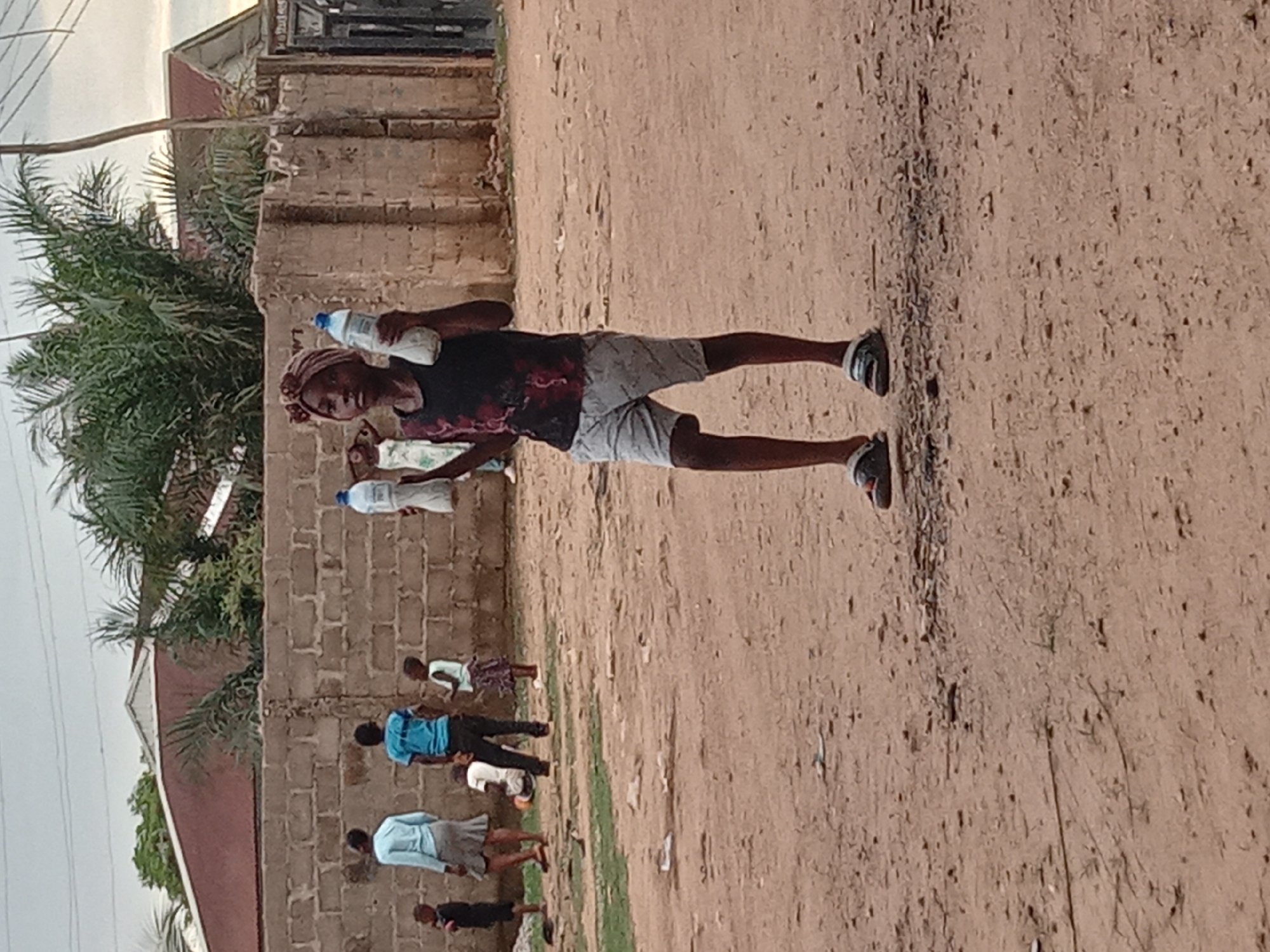 A person is holding two bottles in an outdoor scene on a dirt path in Sabon Tasha, Nigeria. In the background, there's a brick wall, greenery (possibly a palm tree), and several other people standing along the wall. The sky is overcast.