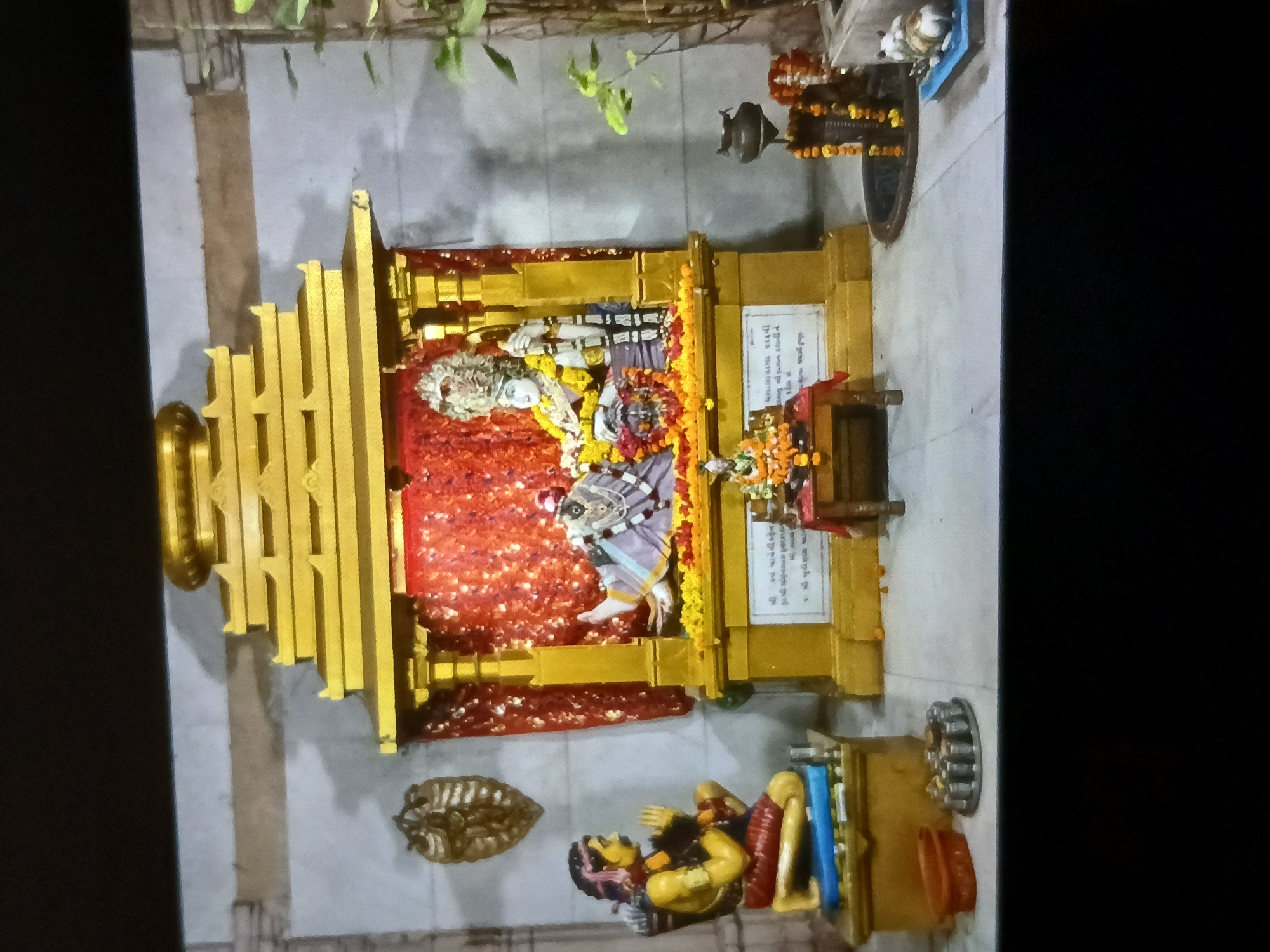 The image shows a Hindu shrine, possibly located in Bengaluru, India. The shrine is predominantly golden, with statues of deities adorned with flowers. Red fabric is draped within the shrine. A small idol rests at the base of the shrine. A sign with script in an Indian language is affixed to the shrine. Additional objects are present, including what appears to be a metal ornament, oil lamps, and prayer books. Plants are visible in the background.
