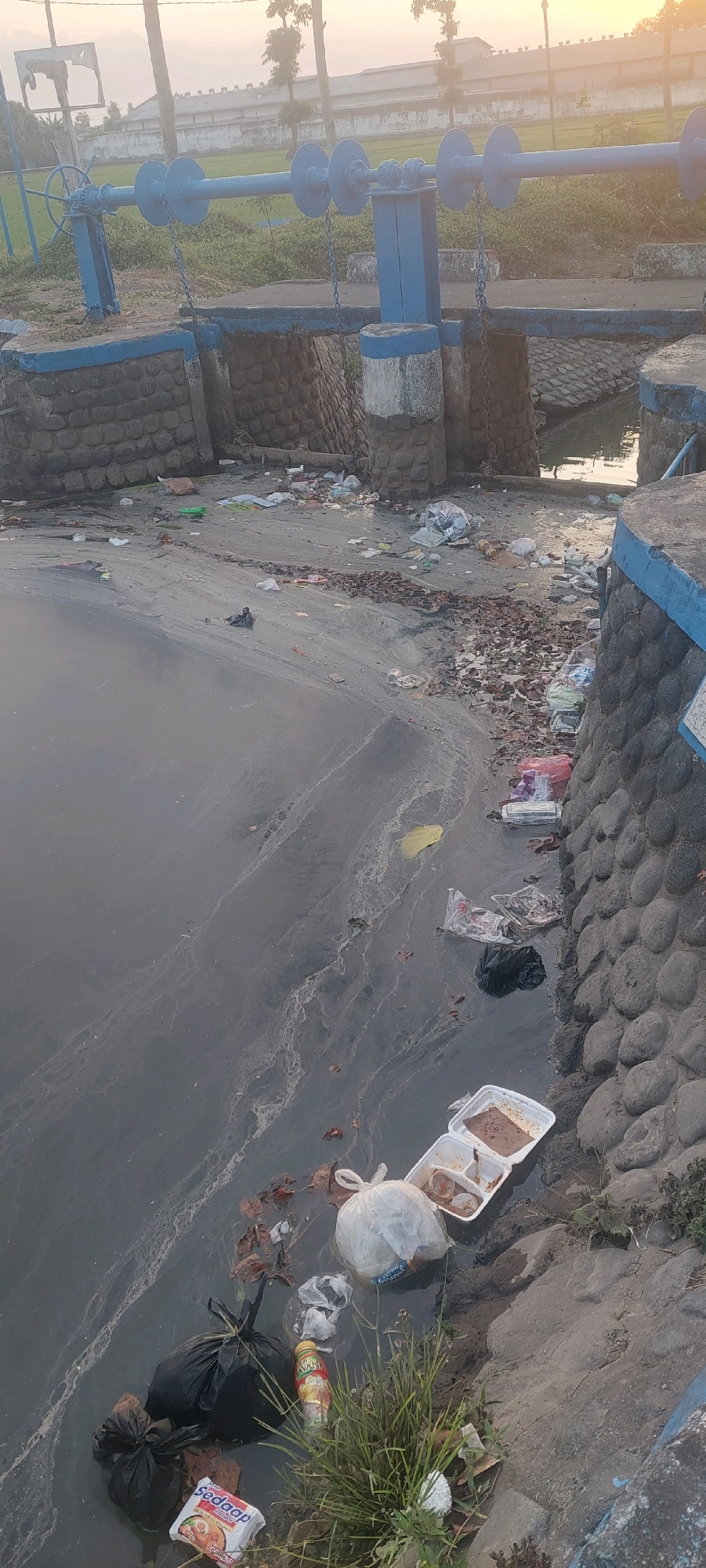 In Jogoyudan, Indonesia, a body of water is heavily polluted with a significant amount of trash, including plastic bags, food containers, and bottles. The water's surface is discolored and shows signs of scum or foam. Along the bank, more debris is visible, interspersed with patches of dry leaves and grass. In the background, there are structures that appear to be part of a water control system, painted blue, with chains and a wheel mechanism. Beyond that, there are buildings and trees, bathed in the soft light of what appears to be either sunrise or sunset. No people are present in the image.