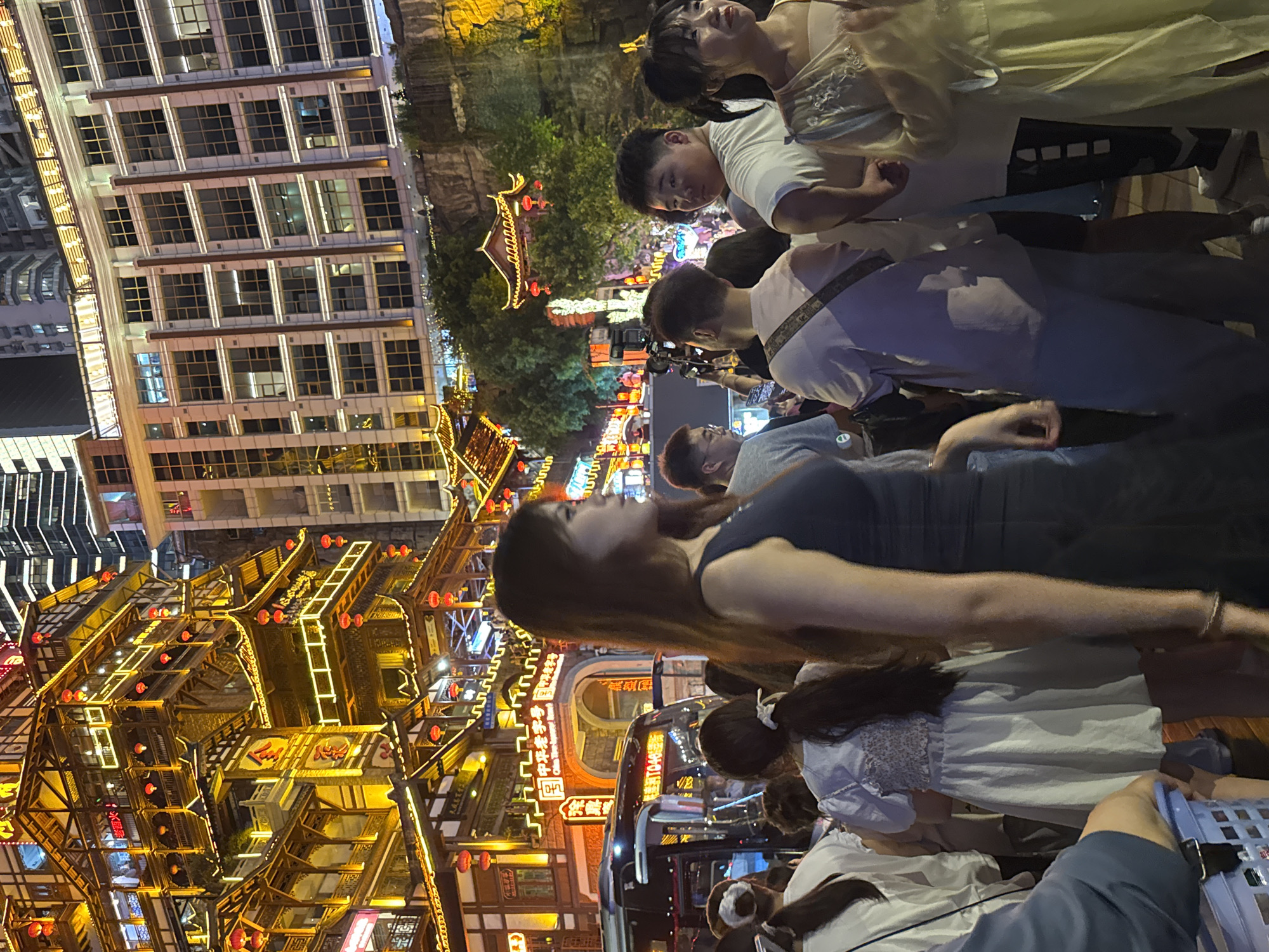 This image captures a lively street scene at night in Yuzhong District, China, likely in an area known for its traditional architecture and vibrant atmosphere. In the foreground, a group of people are gathered, with a woman in a dark sleeveless dress prominently featured in the right-center, looking to her left. To her right, a young woman in a light-colored, possibly embroidered dress is visible. Further to the left, several individuals are seen from behind, including a woman in a white top and another in a blue dress with a patterned bodice. A bus with illuminated signage is parked on the left side of the frame.
The background is dominated by multi-story buildings with distinct architectural styles. On the left, a towering structure with traditional Chinese-style roofing, adorned with red lanterns and illuminated windows, is a prominent feature. To its right, a more modern building with numerous windows and white vertical accents stands tall. Further in the background, the cityscape is visible with more buildings and lighting. The overall environment suggests a popular tourist or entertainment district, with a mix of traditional and modern urban elements. The lighting is predominantly warm, from the illuminated buildings and lanterns, creating a festive and energetic ambiance.