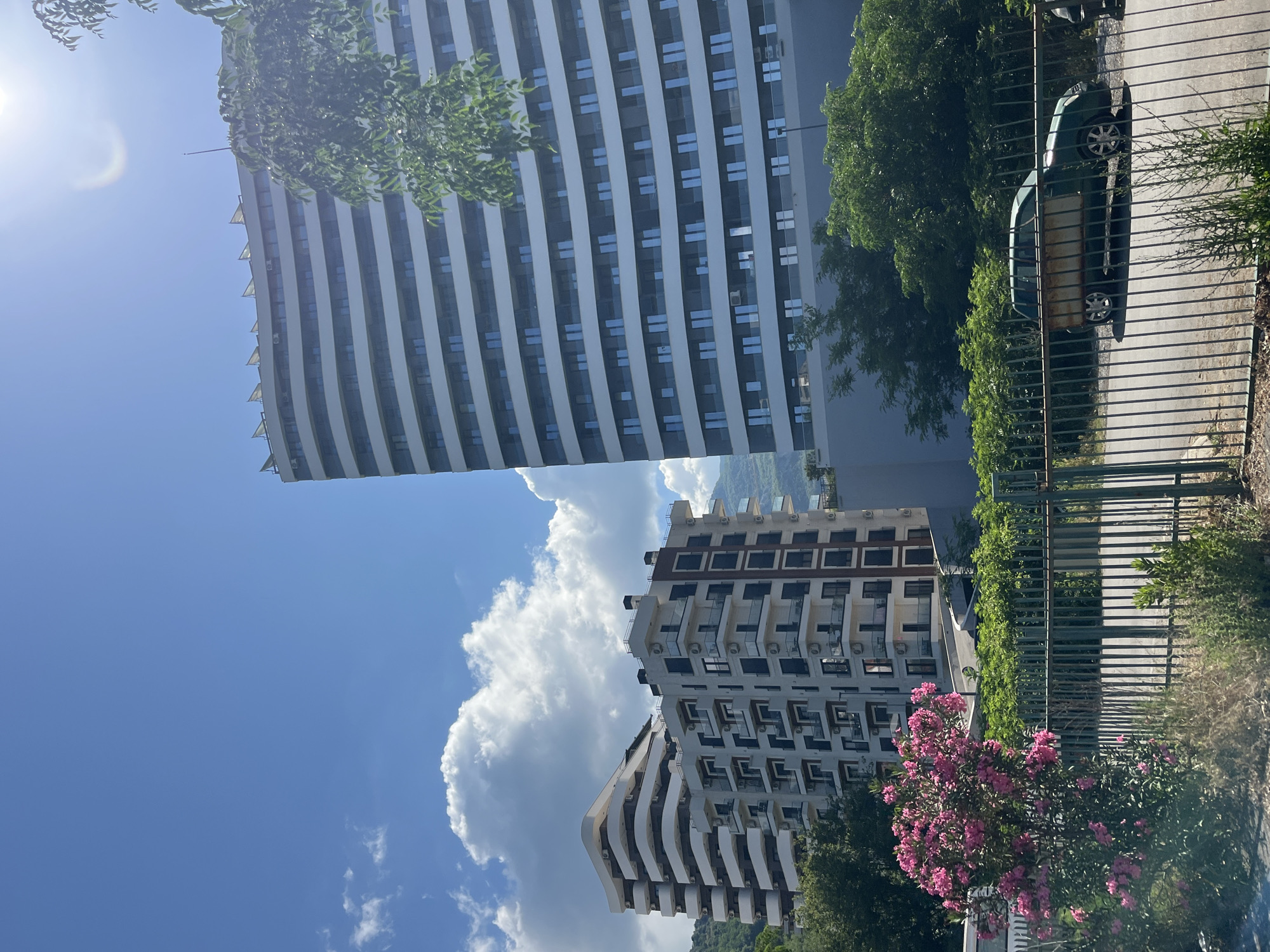 A brightly lit, sunny outdoor scene captured in Boreti, Montenegro, showcases modern architecture amidst lush Mediterranean vegetation. The primary subjects are two contemporary multi-story apartment buildings. The larger building on the left features a curved facade with prominent vertical white bands and numerous blue-tinted windows, some with small triangular sunshades or balconies. The smaller building towards the center-right is more angular, with multiple balconies and dark-framed windows, and visible air conditioning units on its exterior. Another, smaller, similar building is partially visible below it.
The sky is a clear, vibrant blue with several fluffy white cumulus clouds, particularly dense between the buildings. The sun shines brightly from the upper left, creating a noticeable lens flare. Green trees with dense foliage are abundant, bordering the buildings and extending into the background. In the lower right foreground, a large bush with numerous vibrant pink flowers, likely oleander, adds a splash of color.
A green metal fence with vertical bars runs across the right side of the foreground. Behind this fence, a dark green car, possibly a hatchback or small SUV, is parked on what appears to be a paved area. The car's interior or cargo area shows a distinctive brown, perhaps wooden, object. In the distant background, mountainous terrain is faintly visible under the bright sky.
The image depicts a static scene with no discernible human activity. The clear sky and strong sunlight suggest it is daytime, likely mid-morning or afternoon. No visible text is present in the image.
