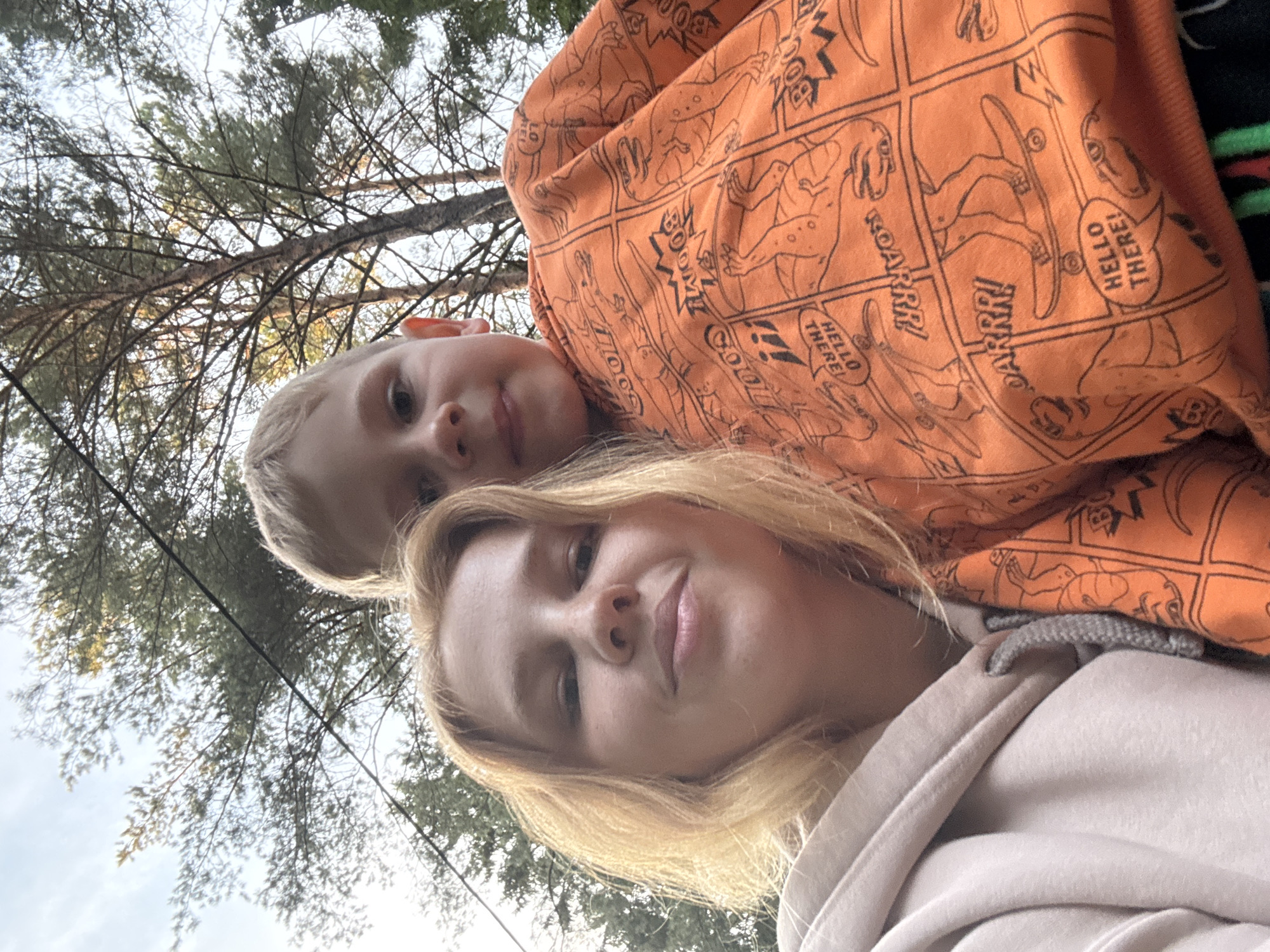 In this low-angle, close-up selfie taken outdoors, two individuals are visible against a backdrop of trees and sky. The person in the foreground on the left is a young woman with blonde hair and fair skin, wearing a light pink hooded sweatshirt. She is looking directly at the camera with a slight smile. To her right and slightly behind her is a young boy, also with blonde hair, wearing a bright orange hooded sweatshirt adorned with a comic-style dinosaur print. He is also looking towards the camera. The image appears to have been taken in a natural, wooded setting, possibly a park or forest, with tree branches and foliage filling the upper portion of the frame. The lighting suggests it is daytime, with the sun filtering through the trees. No specific landmarks or features indicating the location in Anteplevo (Okhotinskoe S/P), Russia, are discernible from the visual information alone.