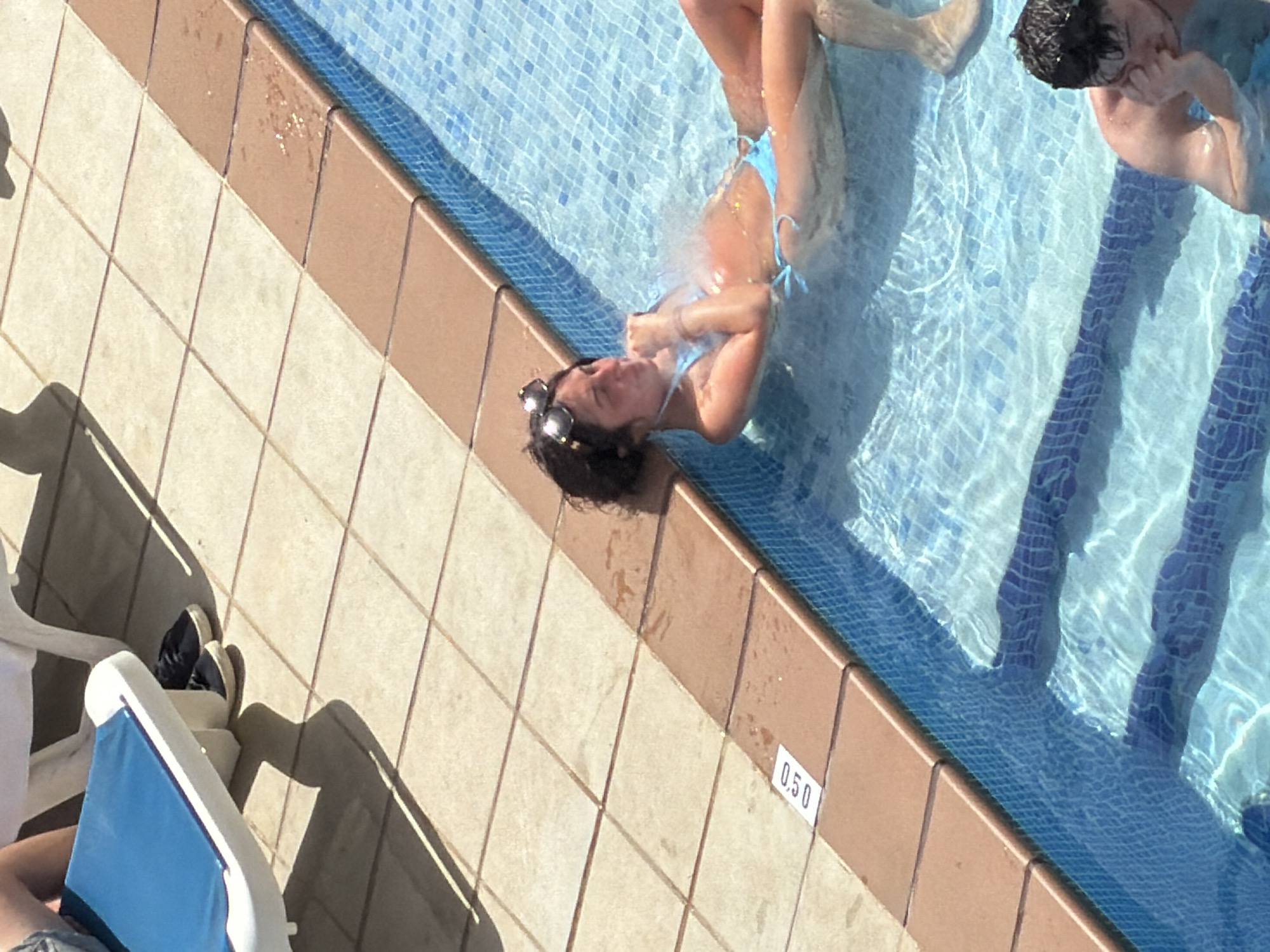 A high-angle shot shows a woman in a blue bikini relaxing in a swimming pool in Sant Antoni de Portmany, Spain. She is lying on her back with her head propped up on the pool's edge, her eyes are closed, and she is holding her hand to her chest. She is wearing sunglasses on top of her head. Another person's legs are visible in the upper left corner of the frame, resting on a white lounge chair. Another person, a male, is partially visible in the lower right of the frame, submerged in the pool, with his hand on his face. The pool has blue mosaic tiles. The edge of the pool is a brown tile border, and beyond that, a tiled patio with light-colored square tiles. A white label with "0.50" printed on it is visible on the patio tiles near the pool's edge, indicating the depth of the pool. The scene is bathed in bright sunlight, casting shadows on the patio.