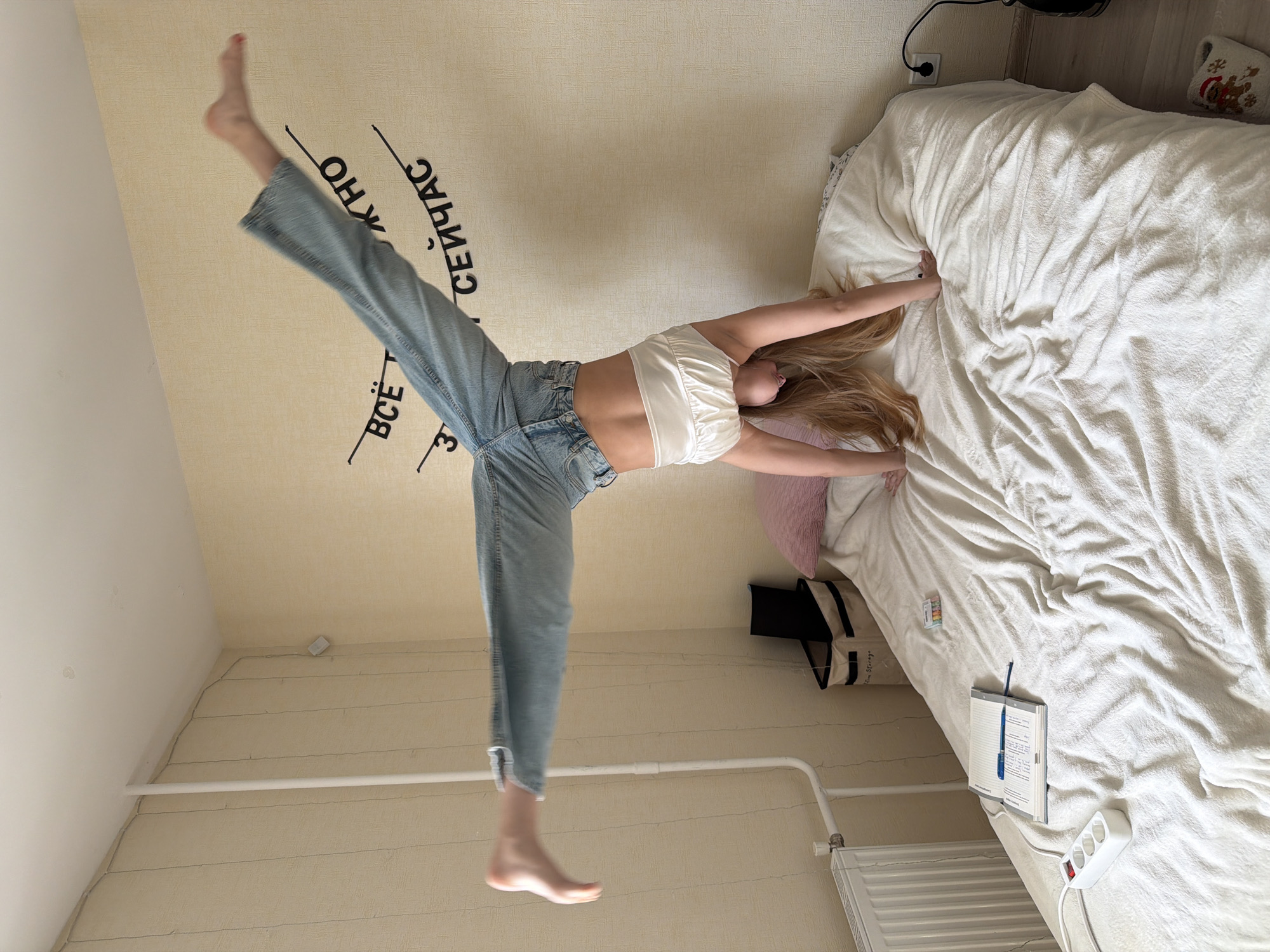 A woman is positioned upside down with her hands on a bed covered with a white comforter. She is wearing a white crop top and blue jeans. Her bare feet are in the air. The wall is light beige with black Russian text near the ceiling. A white baseboard heater is visible along the wall near the floor. There is an outlet with a power strip plugged in. A notebook and pen are on the bed. Based on the available information, the location is Izhevsk, Russia.
