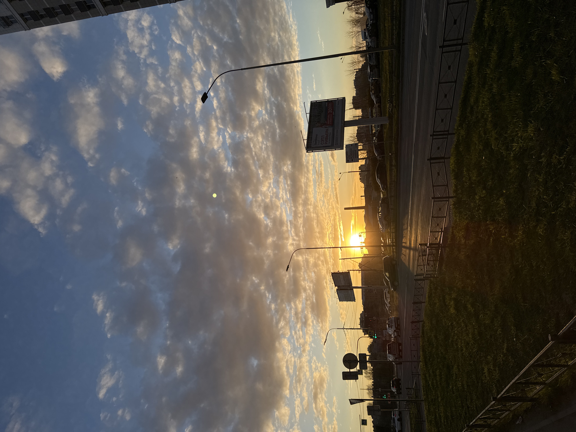 The image shows a cityscape at sunset in Saint Petersburg, Russia. The sky is filled with clouds, illuminated by the setting sun. A road runs through the middle ground, flanked by green vegetation on one side and a fence on the other. Street lamps and billboards line the road. Traffic lights can be seen in the distance. Buildings are visible in the background, silhouetted against the bright sky. There are cars on the road.
