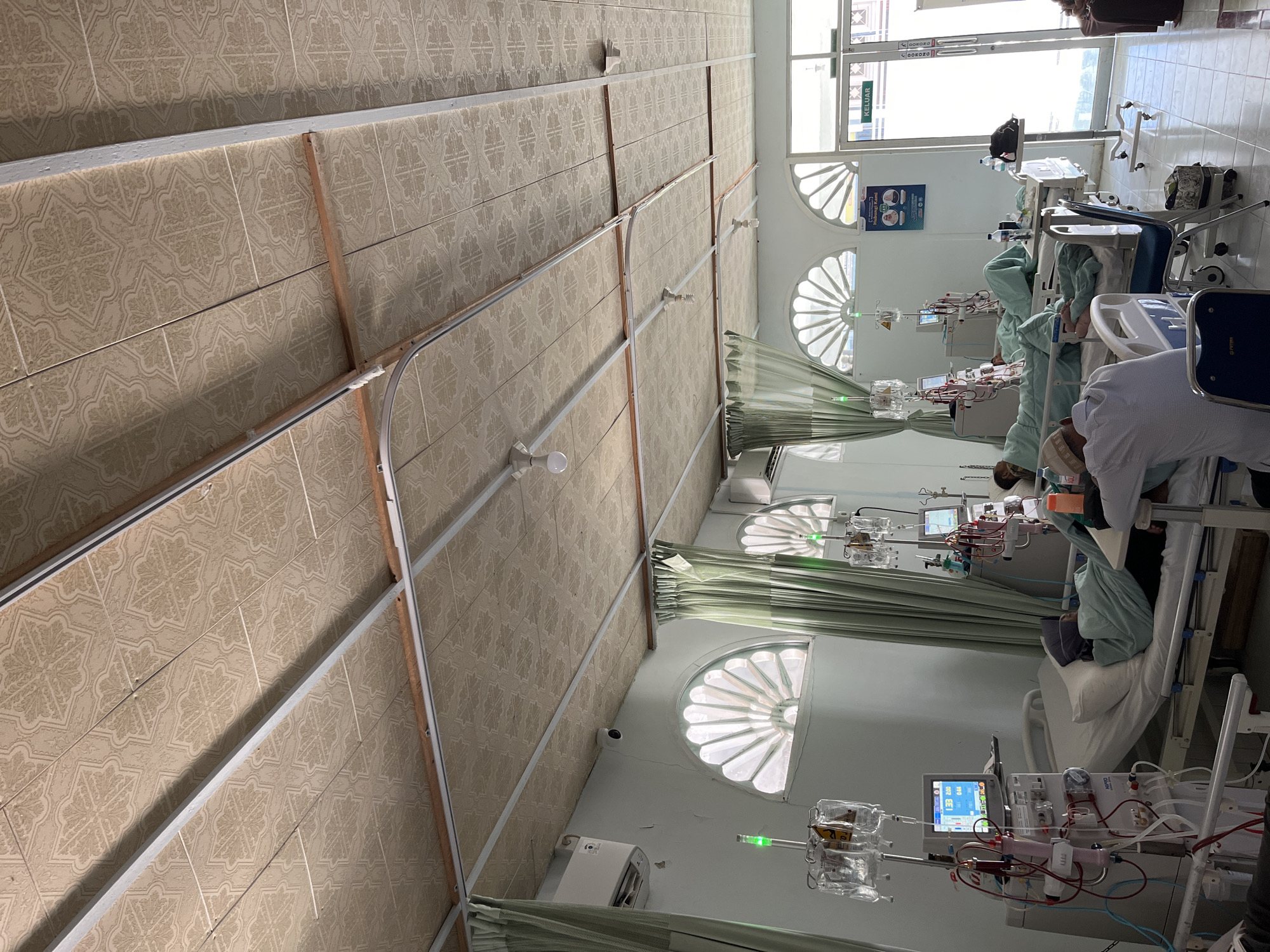 This image provides a detailed view of a medical treatment room, identified as a dialysis center, located in Lhokseumawe, Indonesia. The room features a suspended ceiling constructed from light-toned, patterned square tiles supported by a visible metal framework, with exposed electrical conduit and several ceiling-mounted light fixtures, some with bare bulbs.
The walls are painted a light, neutral color, exhibiting minor surface imperfections in some areas. Distinctive semi-circular, fan-shaped windows, each with multiple panes, are visible along the far wall and partially covered by light green curtains. High on the walls, at least two wall-mounted air conditioning units are present, alongside a small, dome-shaped security camera.
In the foreground left, a patient lies on an adjustable medical bed, connected to a Nipro dialysis machine. The machine's monitor displays numerical data (e.g., "133 280 / 044"). An IV pole with multiple clear fluid bags and a green indicator light stands adjacent to the bed. Two additional patient stations, each comprising a bed, a patient undergoing dialysis, a machine, and an IV pole, are visible further into the room.
In the mid-ground right, a person wearing a white shirt and a peci (traditional cap) is seated on a blue chair beside a patient's bed, facing away from the camera, with their arm resting on the bed. Another individual is partially visible near the room's exit.
A poster on the wall near the exit displays text, including "Hubungi Kami" (Contact Us) and the number "221". The exit itself is a double glass door with a prominent green sign that reads "KELUAR" (Exit). The floor is covered with light-colored square tiles. The room is brightly lit by a combination of natural light entering through the windows and artificial light from the ceiling fixtures, presenting a functional clinical environment.
