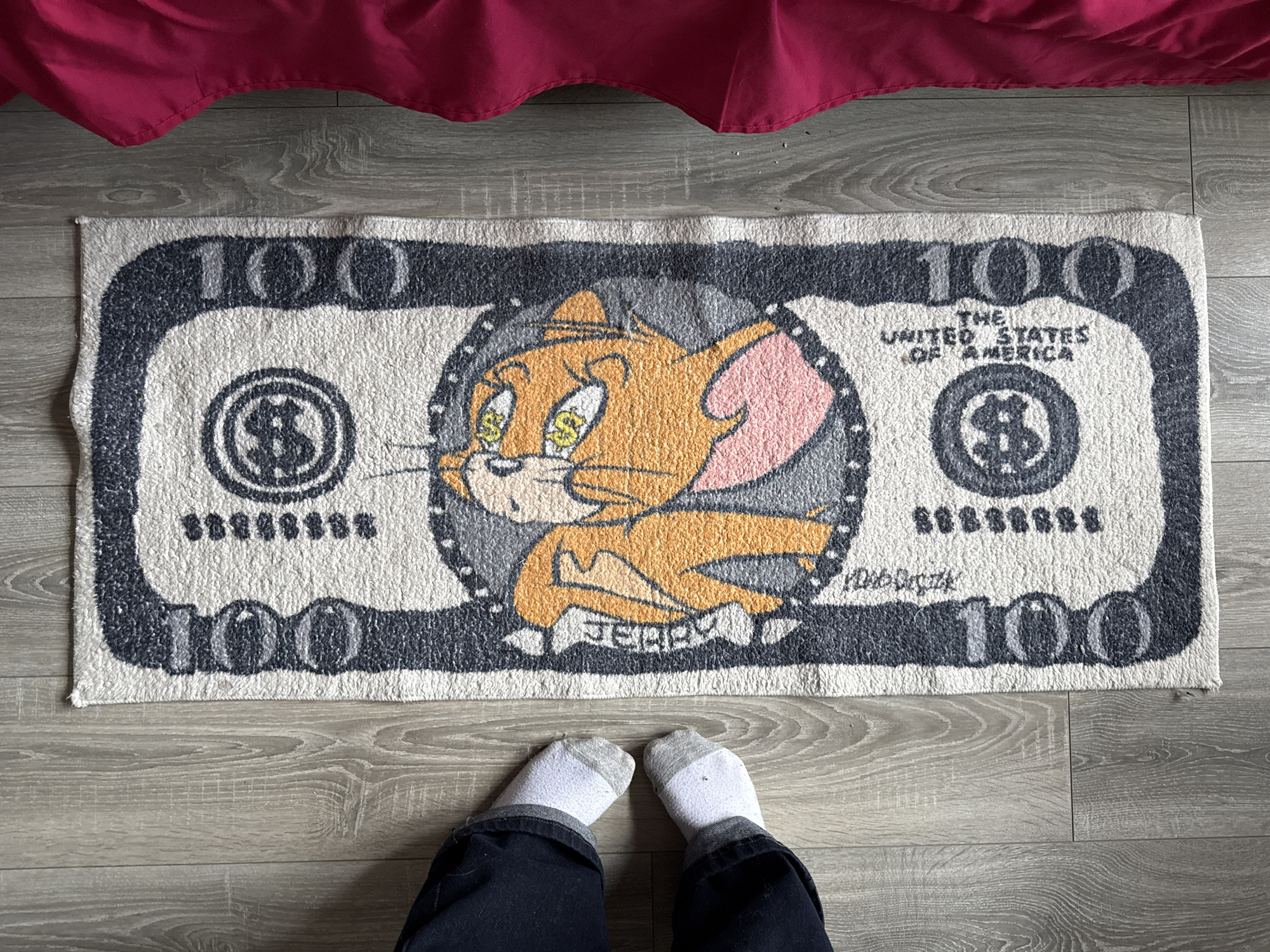 A top-down indoor shot captures a person's lower legs and feet positioned before a distinctive rug laid on a light wooden or laminate floor. The central subject is a rectangular rug designed to resemble an oversized 100-dollar bill.
The rug features an off-white background with bold black and grey outlines and text, mimicking the design of currency. In the center, the cartoon character Jerry, from Tom and Jerry, is prominently displayed in shades of orange, yellow, and pink. Jerry is shown with dollar signs in his eyes, facing towards the left side of the rug, and the word "JERRY" is printed in black text below him. A handwritten signature, possibly an artist's name, is visible on the right side of the central design.
The number "100" appears in large black and grey font in all four corners of the rug, as well as along the top and bottom borders. The text "THE UNITED STATES OF AMERICA" is printed on the upper right side of the bill design. Dollar symbols ("$") are incorporated within circular emblems on the left and right sides of the rug, and a series of repeated dollar symbols are present below "THE UNITED STATES OF AMERICA."
At the bottom of the frame, a person stands on the floor, wearing dark blue trousers with visible cuffs and white socks with grey toes and heels. The floor exhibits a subtle wood grain pattern. At the very top of the image, a dark red or maroon fabric, possibly a bed skirt or curtain, drapes into the frame. The scene is set indoors, with the user specifying the location as Perm, Russia.