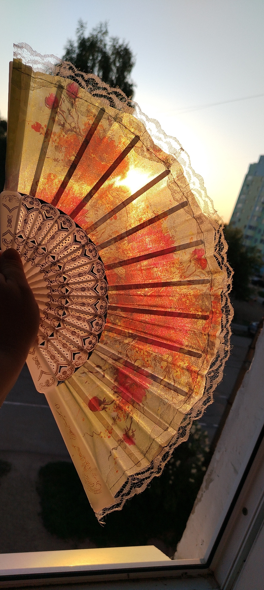 A hand holds an open decorative fan with an orange floral pattern against the light of a sunset. The fan has a lacy trim and the light shines through the translucent fabric. A building and some trees can be seen through the fan in the background. The location is Chaikovsky, Russia. The fan is being held indoors, next to a window.