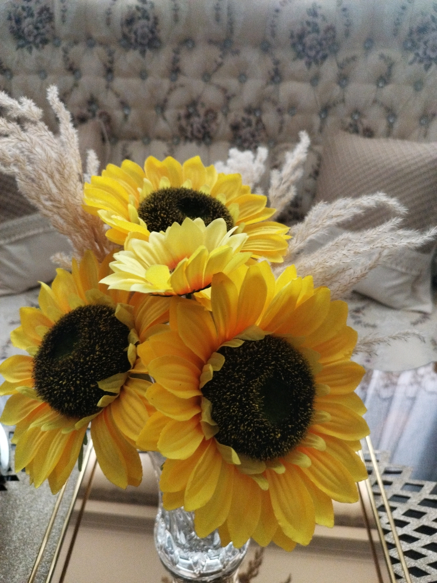 The image shows a close-up of a floral arrangement featuring several artificial sunflowers and dried pampas grass. The sunflowers are vibrant yellow with dark brown centers, and they are arranged in a clear glass vase. The pampas grass is a light beige color and adds a wispy texture to the arrangement. In the background, a tufted upholstered headboard or furniture piece with a floral pattern is visible, along with pillows. The overall scene suggests an indoor setting, likely a bedroom or living area, with a decorative focus on the floral display. The location of Eslamshahr, Iran is not discernible from the visual information presented in the image.