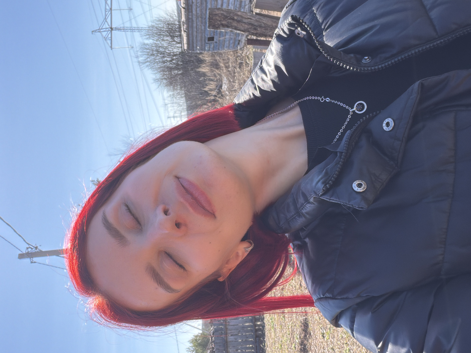 The image is a close-up shot of a woman with red hair, eyes closed, facing upwards towards the sky. She is wearing a black puffer jacket over a black top and a silver necklace. The background shows power lines, bare trees, and a dilapidated wooden structure, suggesting an outdoor location. Based on the prompt, the location is Chaikovsky, Russia, implying an urban or semi-urban setting.