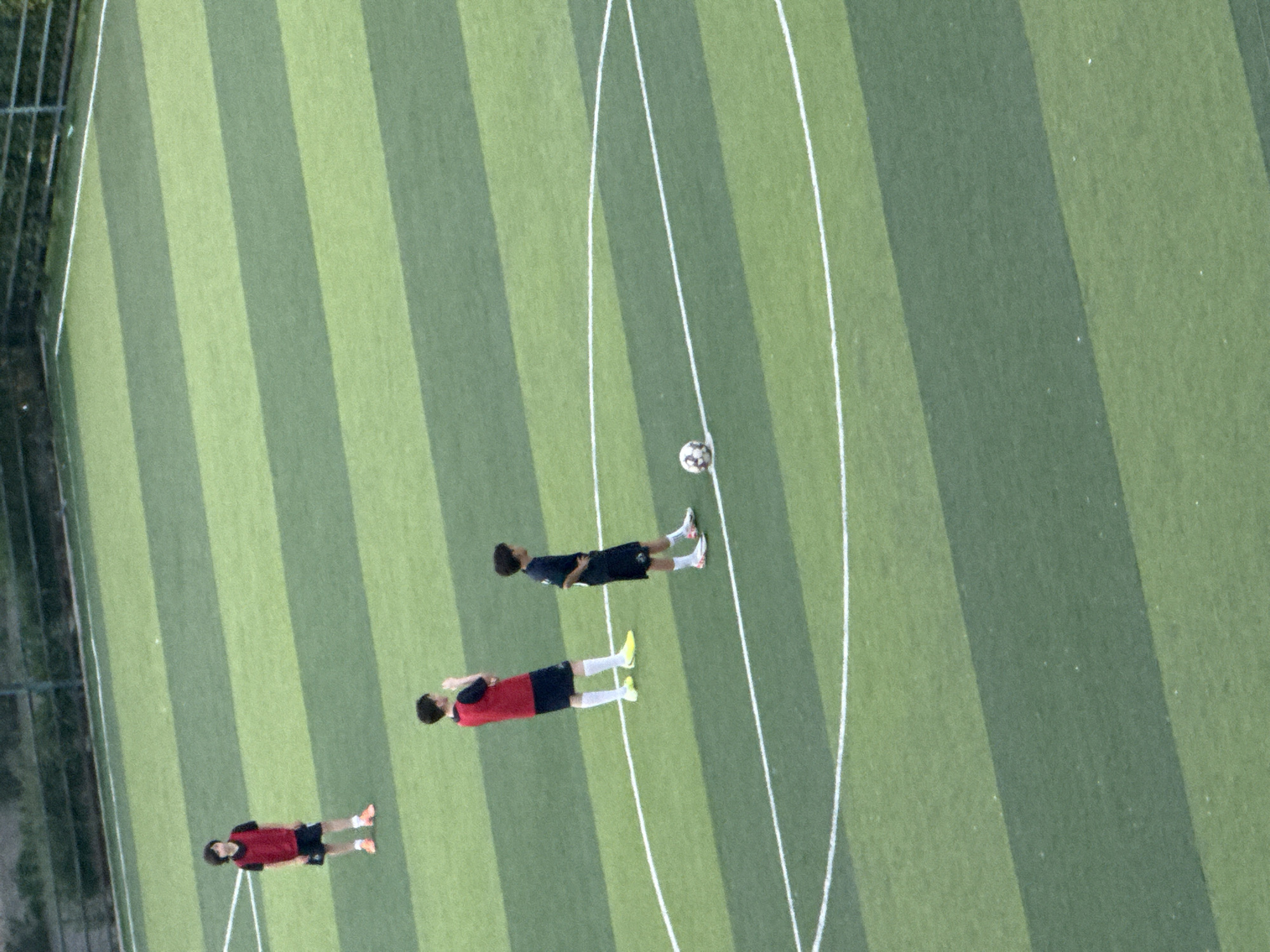An aerial, high-angle, eye-level shot shows a children's soccer practice on an outdoor artificial turf field. The field has alternating stripes of dark green and light green. In the foreground, a child in a dark blue jersey and shorts stands with a soccer ball at their feet. In the middle ground, another child in a red jersey and dark blue shorts stands with their hands behind their back. In the background, a third child in a red jersey and dark blue shorts stands with their hands at their sides. A chain-link fence runs along the top of the frame, behind the field. The weather is overcast, and the lighting is diffused. The image does not contain any identifiable landmarks to suggest its location in Tbilisi, Georgia.