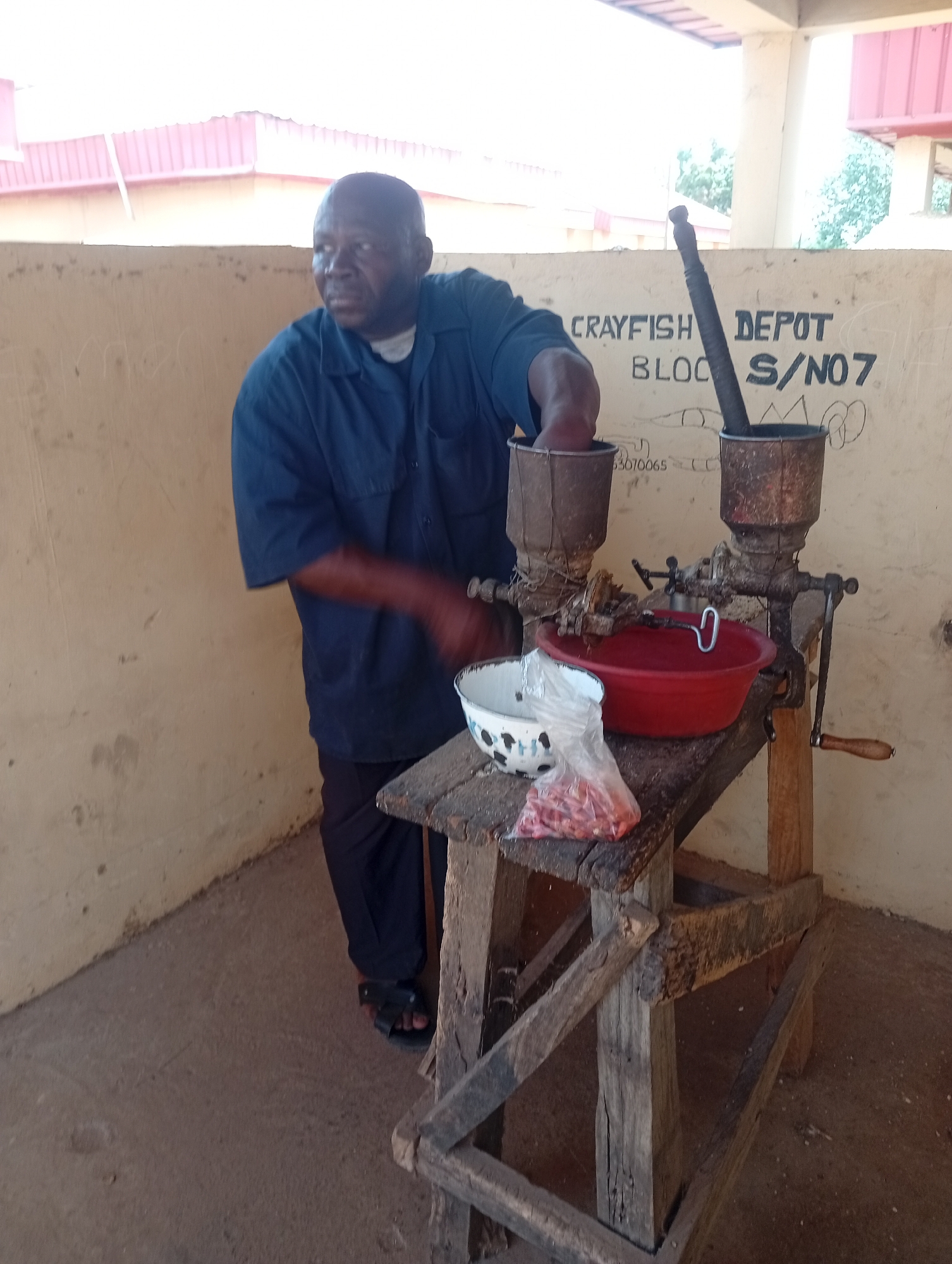 A medium shot depicts a man operating a grinding machine in what appears to be an outdoor setting. The man, dark-skinned and bald, wears a navy-blue button-down shirt and dark pants. He is standing to the left of a worn, wooden table that supports two vintage grinding machines. He is actively using one of the machines, his hand visible near the hopper.
On the table, a red plastic bowl sits between the machines, and a plastic bag filled with red items, possibly peanuts or dried peppers, is placed on the table's edge near a white and blue enamel bowl. The grinding machines are old and rusty, with hand cranks for operation.
The background is a plain, light-colored wall with graffiti or markings, including the words "CRAYFISH DEPOT BLOCK S/NO7". The area appears to be under a partial roof, with glimpses of buildings and greenery visible beyond the wall. The location is Unknown, Unknown.