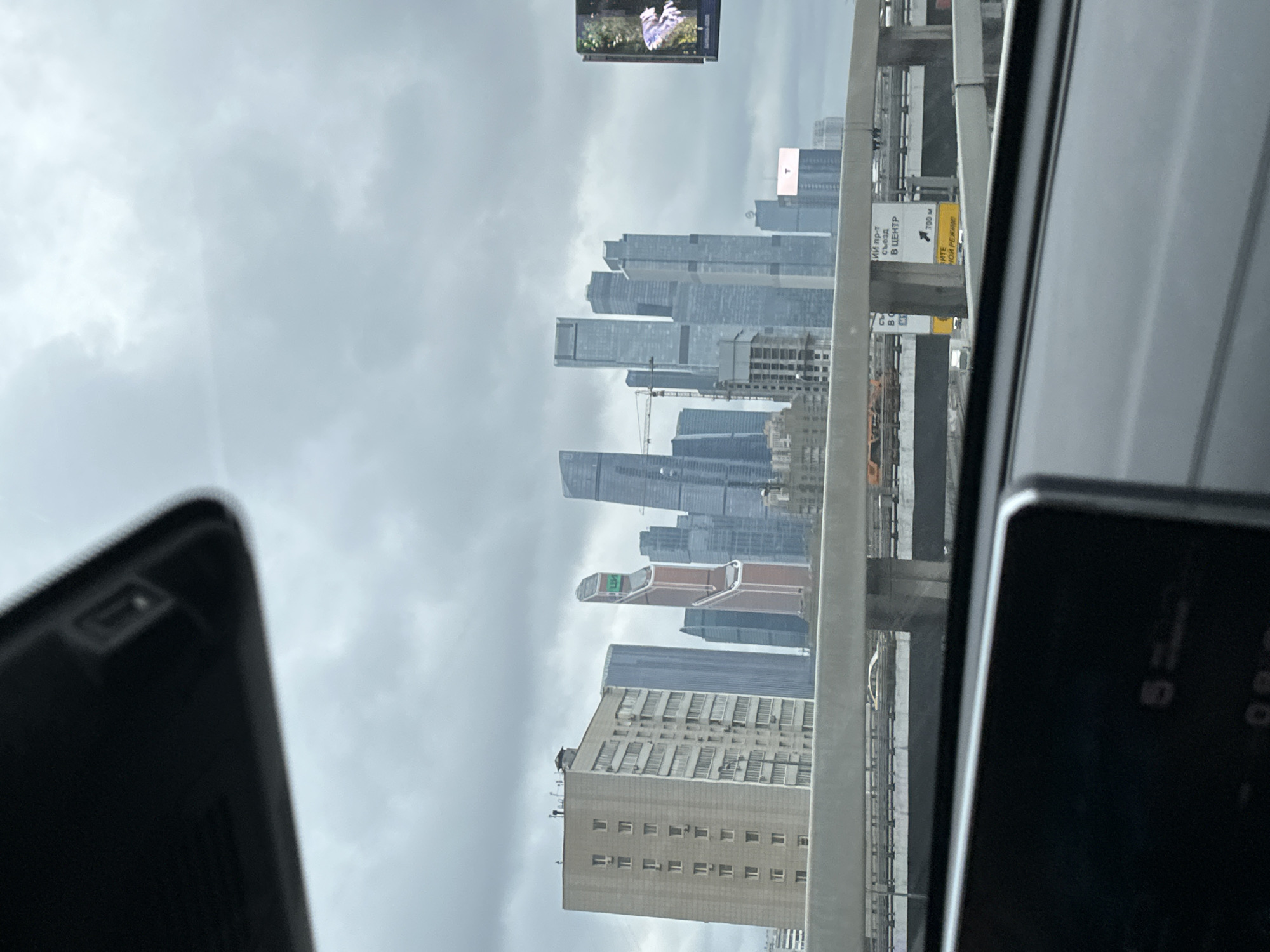 The image captures a view of the Moscow International Business Center (Moscow City) skyline through a car window. High-rise buildings of varying heights and architectural styles dominate the cityscape, shrouded in a hazy atmosphere under a cloudy sky. The lower part of the image includes the upper section of a multi-story building with rows of windows. Road signs and infrastructure elements suggest the scene is taken from a road or overpass. The dashboard of a car is visible in the foreground, partially obscuring the view.