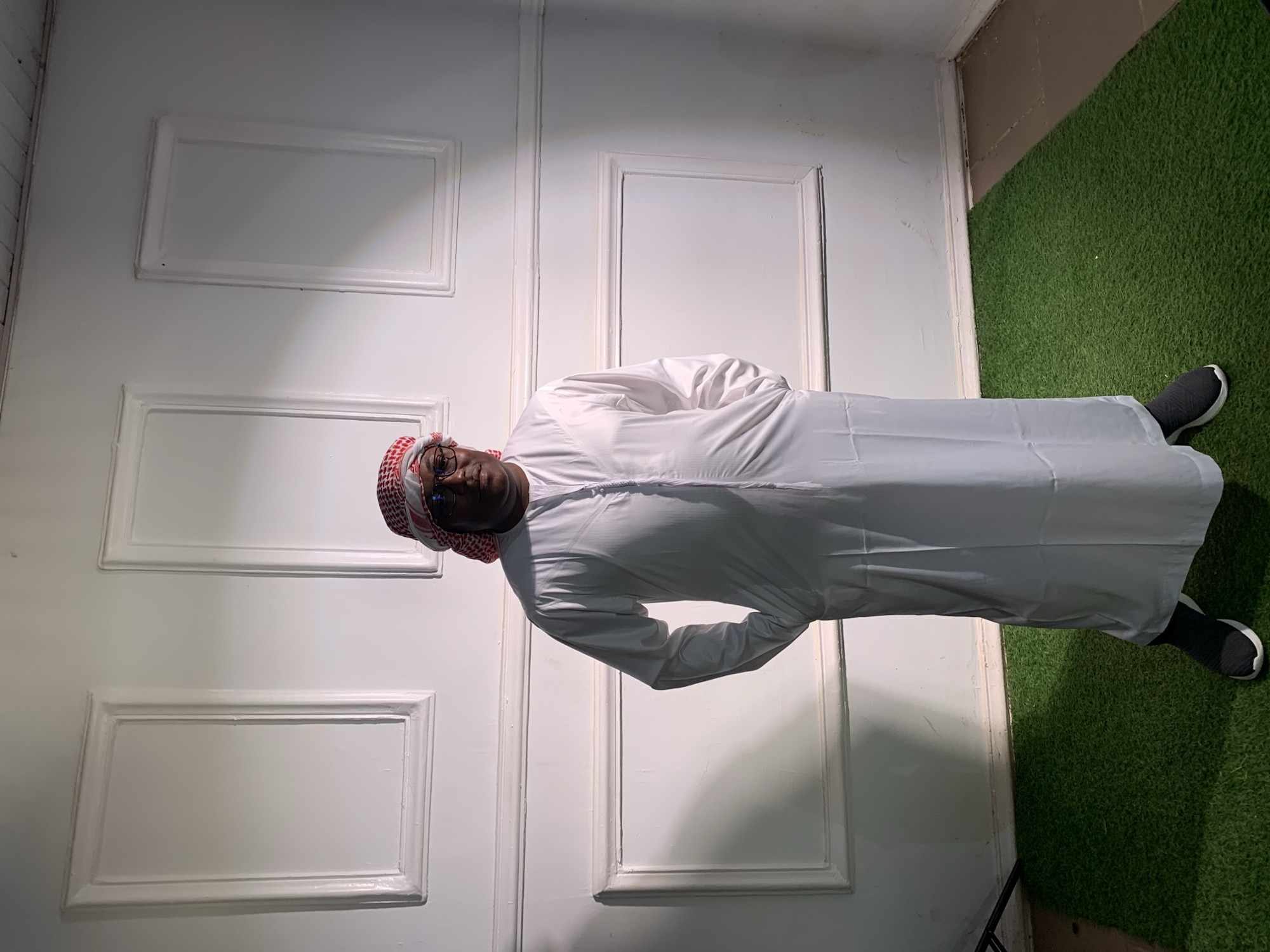 A man stands in a room with a white wall resembling a door with three recessed rectangular panels. He is dressed in a white thobe and a red and white keffiyeh. He wears black and white sneakers. The floor next to the wall is covered with artificial green turf. The location is not identifiable as Barakin Akawo, Nigeria.