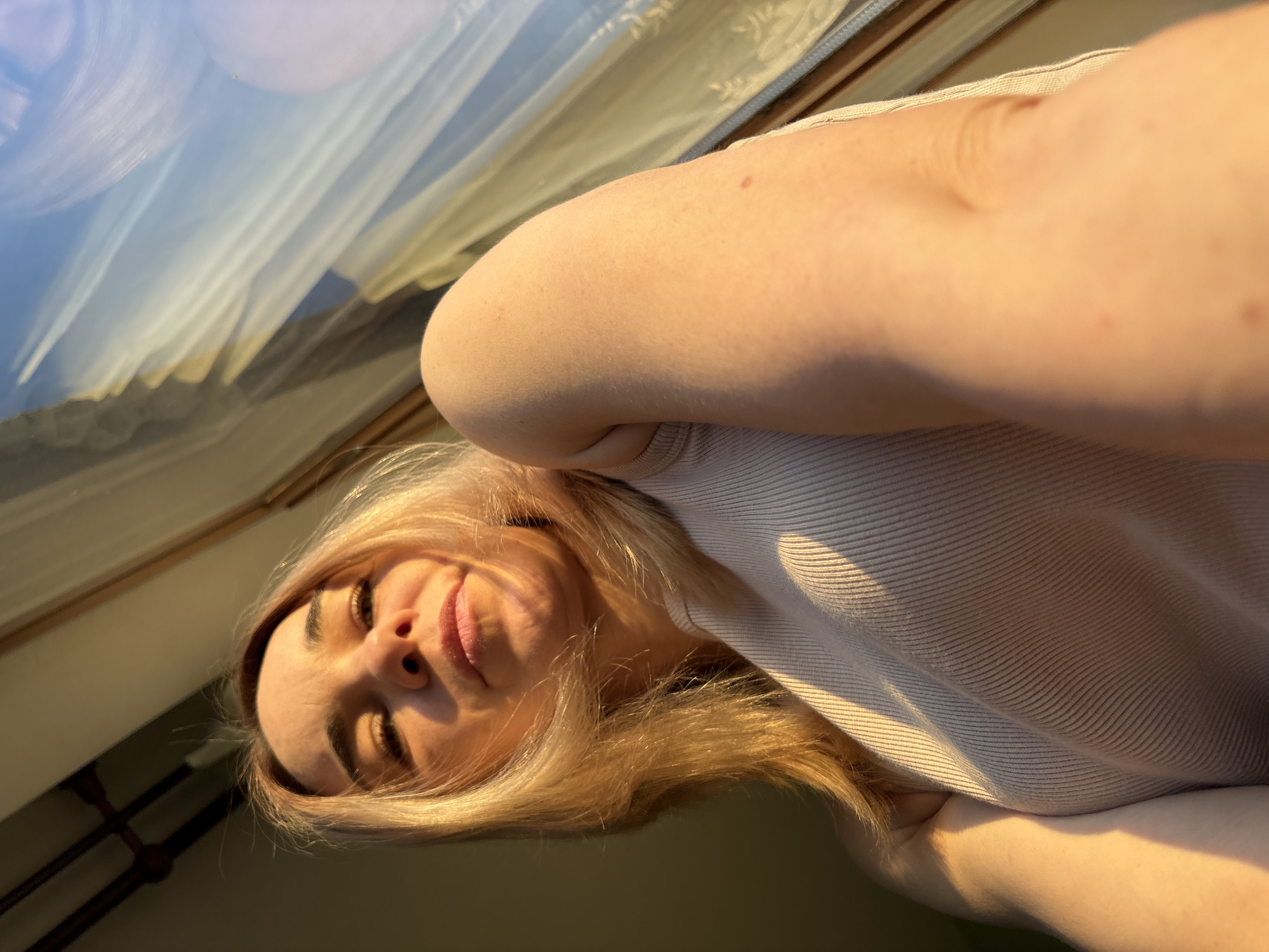 This media file is a close-up, low-angle selfie of a young woman taken indoors. The woman, with blonde hair, fair skin, and dark eyebrows, looks directly at the camera with a slight smile or smirk. Her head is tilted, and her body is partially visible, wearing a light-colored, ribbed short-sleeved top. Her arm extends into the upper right of the frame.
The scene is bathed in warm, golden light, creating distinct shadows on her face, hair, and clothing, characteristic of either early morning or late afternoon golden hour sunshine.
In the upper left background, a large reflective surface, likely a window, shows a blurred reflection. This reflection features soft blue and white tones, suggesting a sky, alongside muted yellow and brown shapes that could be distant landscape features or architectural elements.
The overall impression is casual and personal, captured within an indoor setting near a window. The photo was taken in Sinyavino, Russia. No visible text is present in the image.