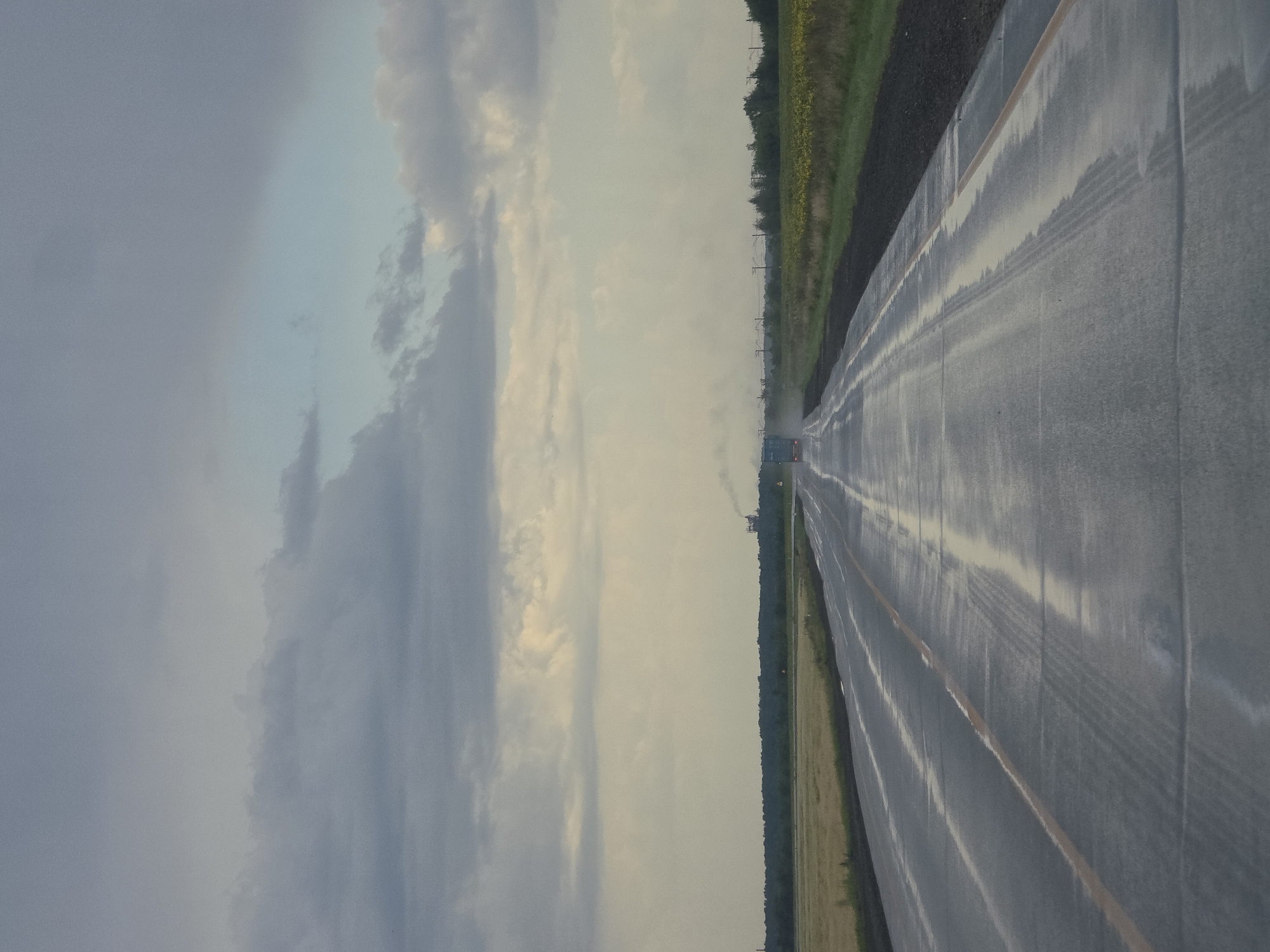 An eye-level, medium shot captures a wet, empty asphalt road stretching towards the horizon under a cloudy sky in Nagorny, Russia. The road is segmented by a yellow line and shows reflections of the sky and surrounding landscape, indicating recent rain. The environment appears rural, with fields of wheat and sunflowers bordering the road. In the distance, a faint industrial structure with a smokestack is visible, as is a single blue truck on the road. Power lines also dot the landscape.