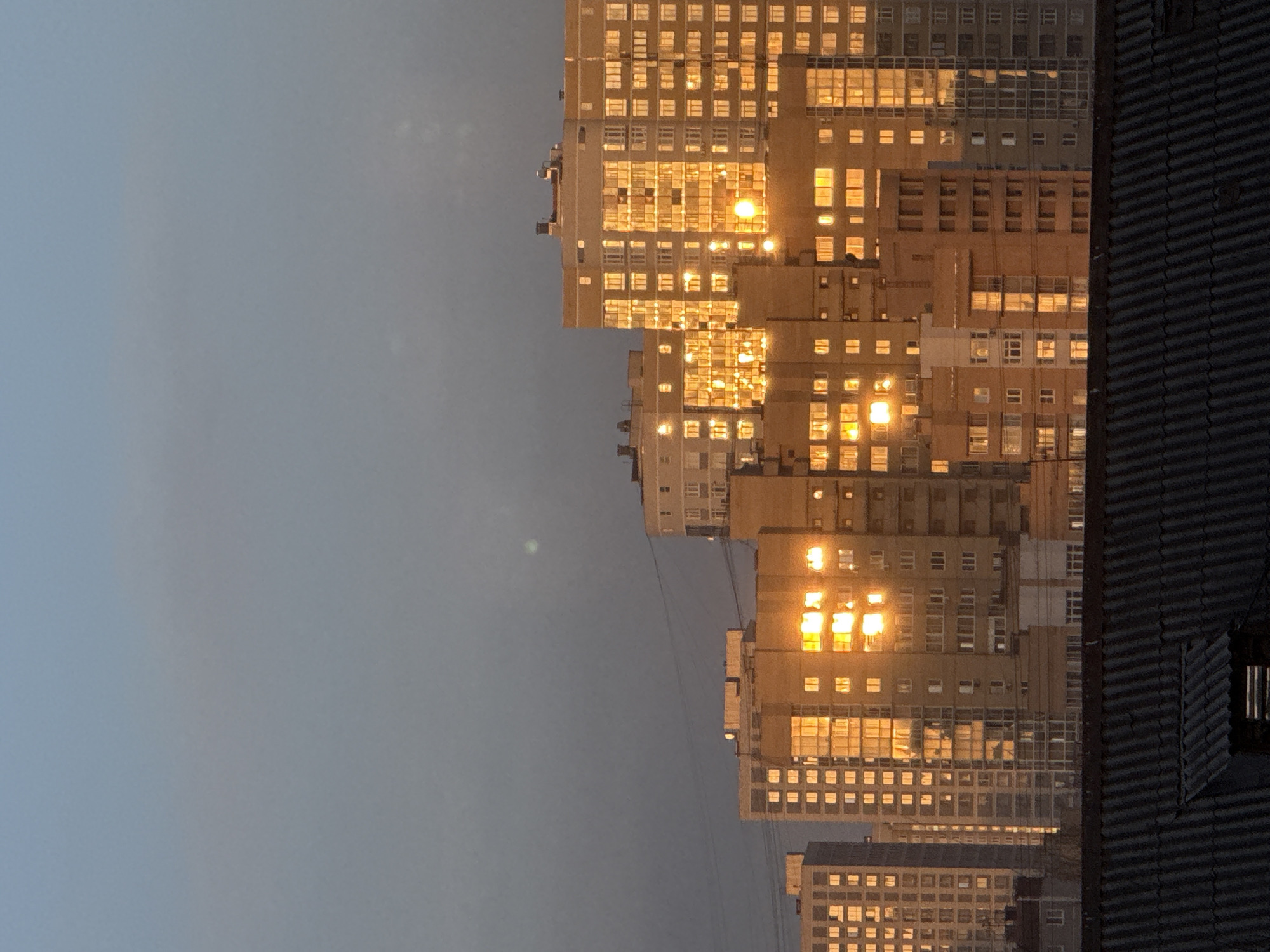 The image shows apartment buildings in Yekaterinburg, Russia, at twilight. The buildings are tall and feature many windows. The windows reflect the light of the setting sun, creating a golden glow. A dark, corrugated structure, possibly a rooftop, is visible on the right side of the frame. The sky is a muted grey-blue. There are no people visible in the image.