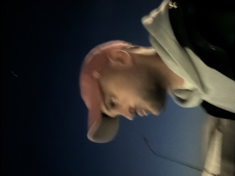 A blurred outdoor image, captured in Sofia, Bulgaria, depicts a male individual in profile. The subject is seen from the chest up, facing the right side of the frame, under an evening or night sky. He is wearing a red baseball cap, a light grey hooded sweatshirt, and a dark outer jacket. The man has facial hair, including a beard and mustache. His gaze is directed forward and slightly upward. He holds a pair of dark-framed glasses by an arm in his left hand, which is partially visible in the lower-left foreground. The background is a dark blue, indicating low light conditions, with a faint, lighter horizon line. No specific landmarks are discernible.