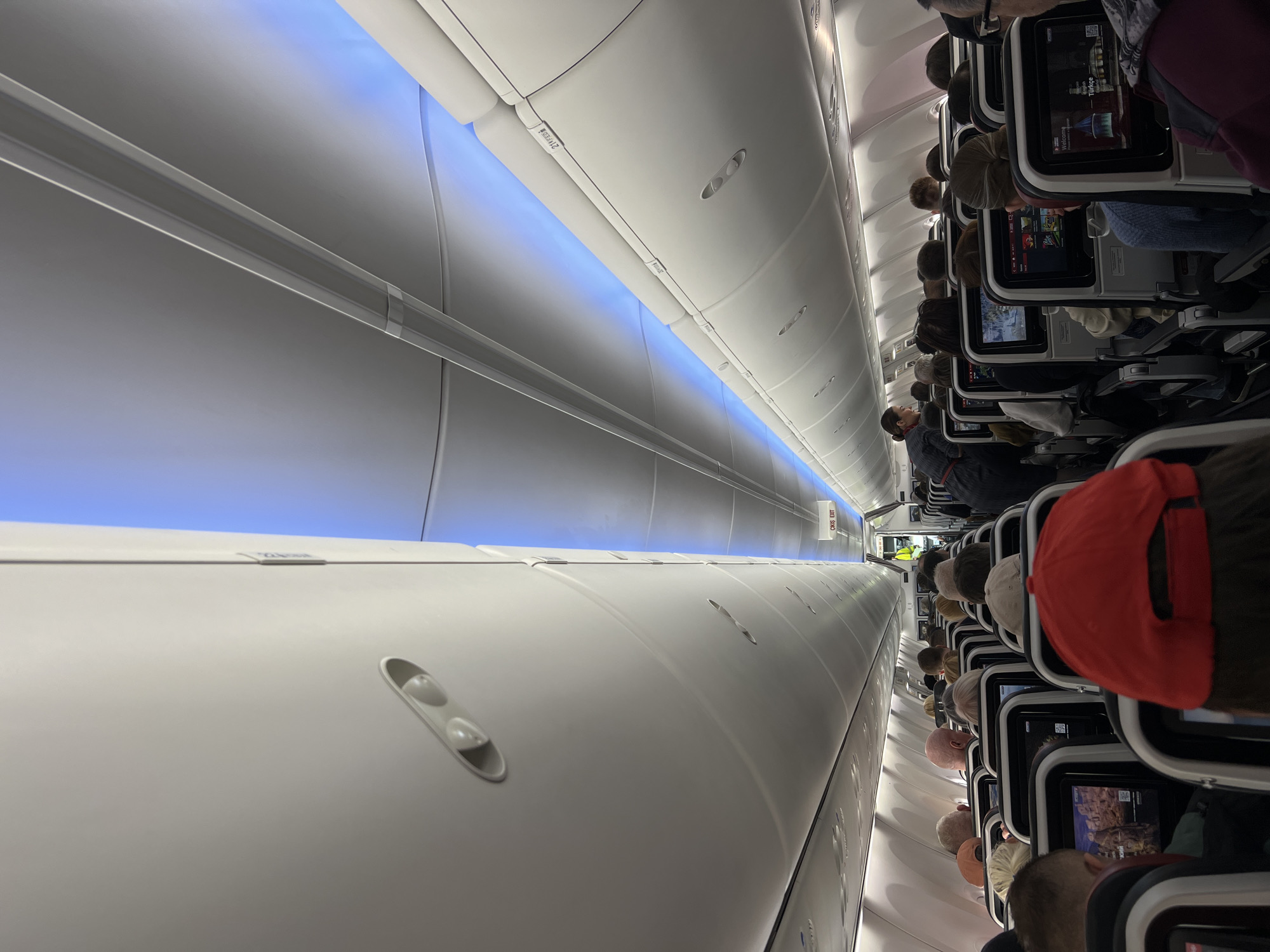 The image captures the interior of a modern airplane cabin, viewed from a slightly elevated, angled perspective. The scene shows rows of passengers seated in what appears to be an economy or premium economy section. Many passengers are visible from behind, their heads facing forward towards seat-back entertainment screens. One person in the immediate foreground wears a red baseball cap, also facing away from the camera.
The cabin features white, sleek overhead compartments running along both sides of the aisle. A striking detail is the ambient blue LED lighting strip that runs the length of the ceiling, casting a soft glow. Overhead panels show individual air vents and reading lights.
Several passengers are actively engaged with their personal seat-back screens, which display various content, including what appears to be movies or television shows. On one screen, the word "Welcome" is visible, along with "Türkçe," suggesting a Turkish airline or a flight with Turkish language options.
Towards the rear of the cabin, a red sign prominently displays "EMERGENCY EXIT." Another sign further back reads "CREW EXIT." The aircraft interior appears well-maintained and modern, indicating a contemporary passenger jet. The overall activity depicted is passengers settled in for a flight, possibly during boarding, cruising, or deplaning, given the presence of people in their seats and the active screens. There are no strong emotional cues, with passengers appearing calm and occupied.