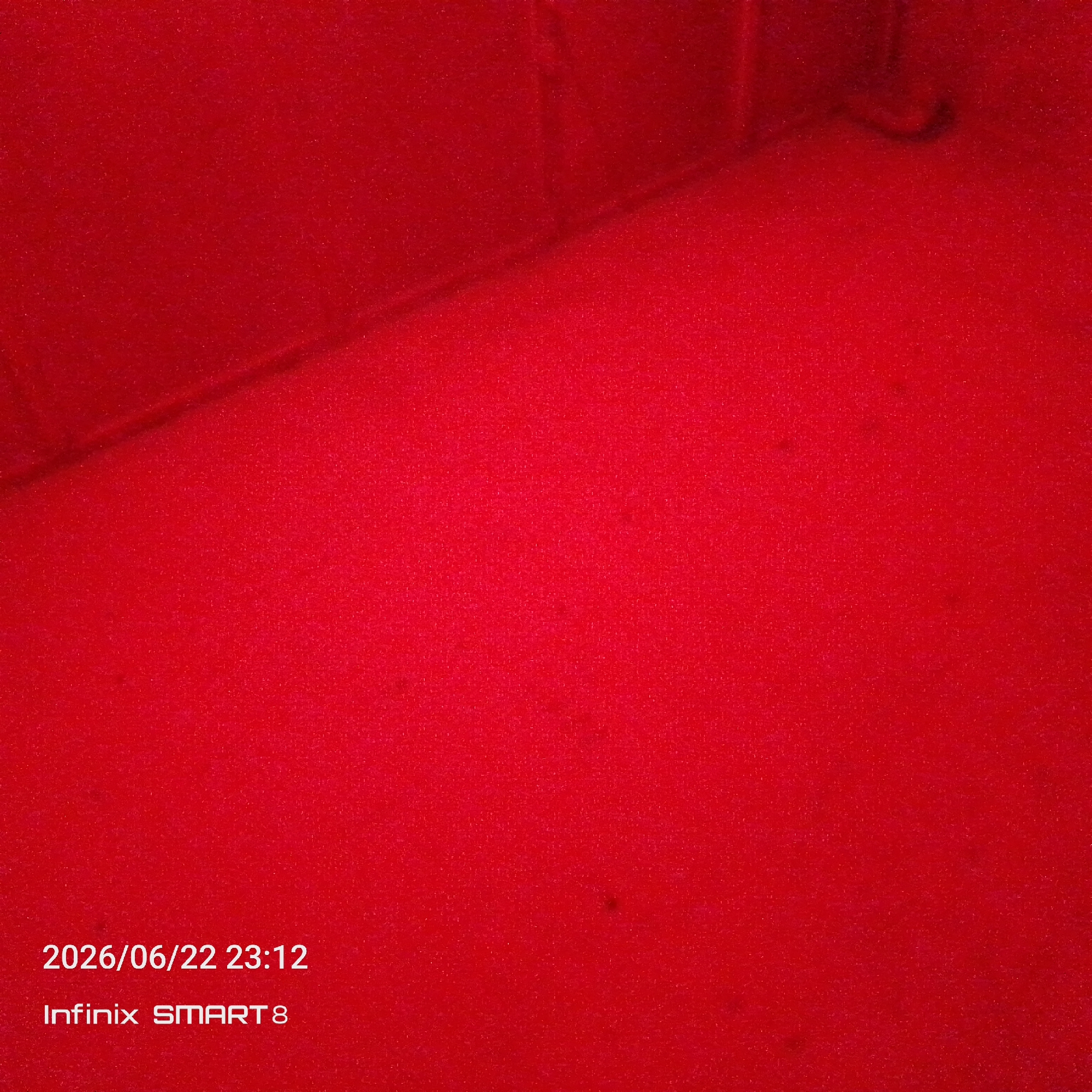 The image displays a close-up view of a textured red surface, possibly carpet or fabric, under red lighting. The texture of the surface is granular and appears to be made up of small fibers. In the upper left quadrant, there are darker shadows and a curved, indistinct shape that suggests the presence of an object or a corner of a room. The lower portion of the image contains white text overlay. The top line of text reads "2026/06/22 23:12", indicating a date and time. The second line of text reads "Infinix SMART 8", which is likely the model of the device used to capture the image. The scene is static and lacks any discernible human presence or activity. The overall impression is that of an indoor setting, possibly a room or a studio, with a dominant red color scheme.
