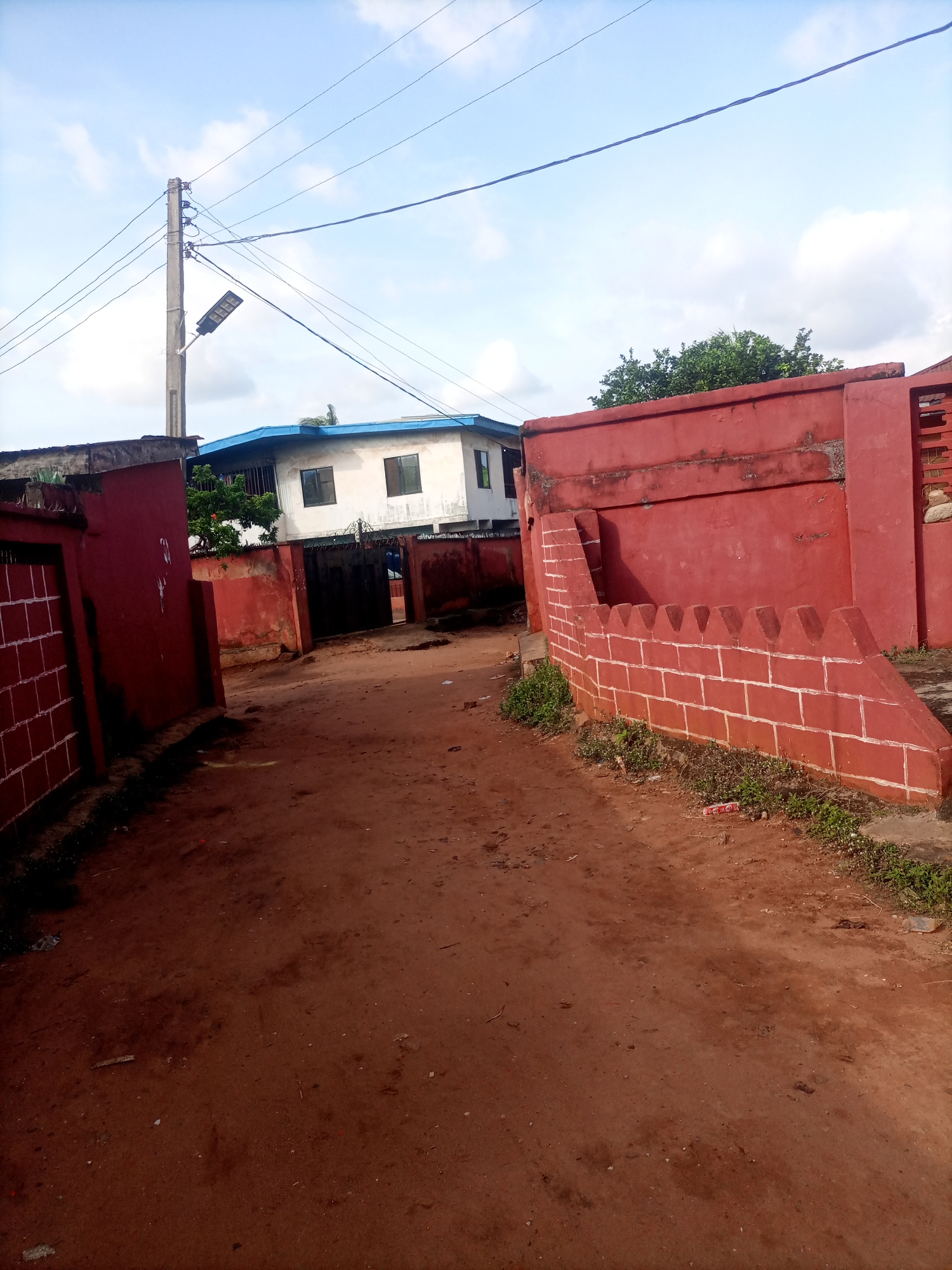 The image shows a street in Nkpor, Nigeria. The street is unpaved and made of red dirt. On the right side of the street is a red brick wall with white painted accents, topped with crenellations. To the left of the street, there is another red wall and a gate with a white grid pattern. Further down the street, there is a white building with a blue roof, and another building partially visible behind it. A utility pole with wires and a solar-powered street light stands on the left side of the image, behind the red wall. The sky is cloudy. There are no people visible in the image. The overall scene depicts a residential or commercial area within the city.